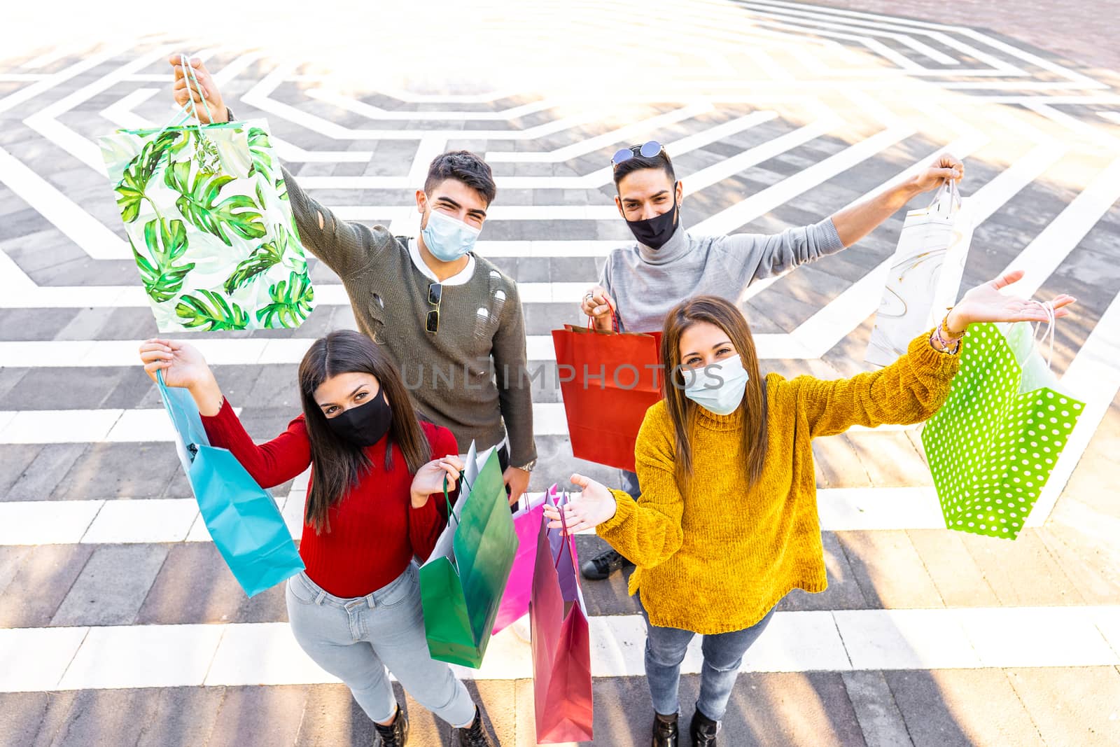 Group of happy Caucasian young friends taken from above wearing Coronavirus face protection medical mask showing their colored shopping bags - Concept of new normal consumer activity in the city by robbyfontanesi