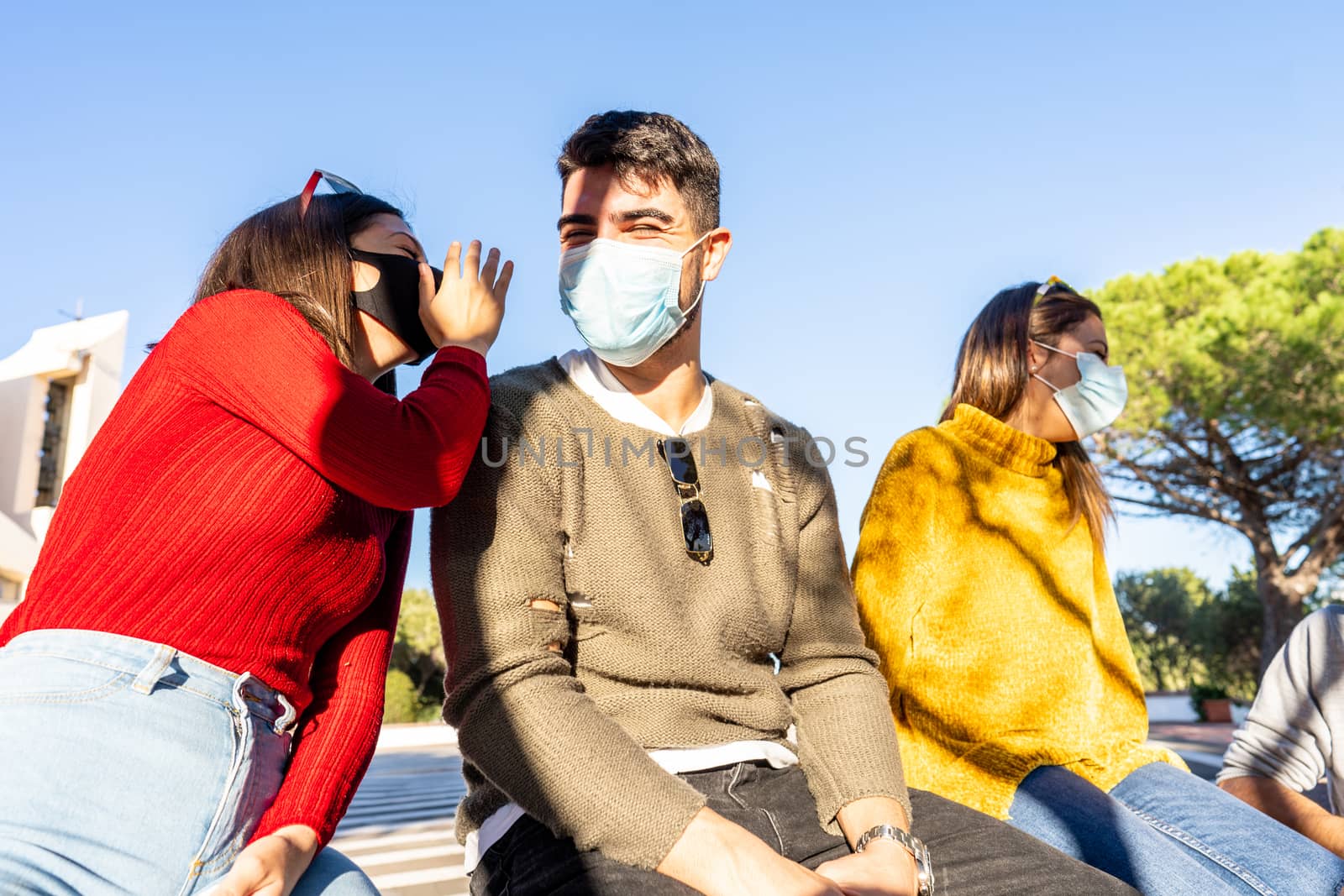 Young Caucasian woman speaks in the ear of her boyfriend sitting on a wall of the city square with friends wearing Coronavirus safe protection medical mask - Carefree couple having fun outdoor by robbyfontanesi