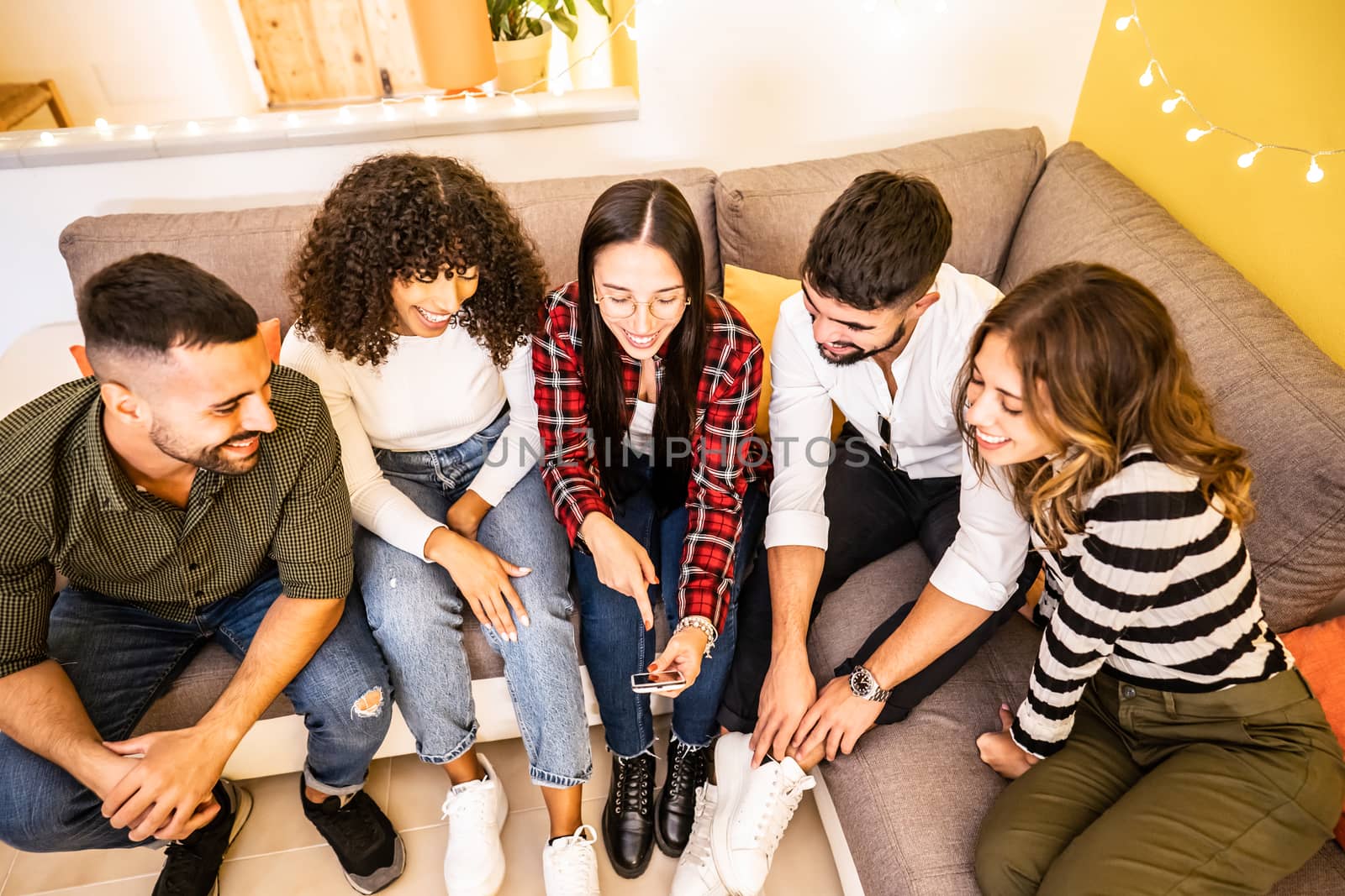Group of mixed race friends having fun at home sitting on the sofa looking at the smartphone - Multiracial young people using social network to share happy moments online - Woman pointing her phone