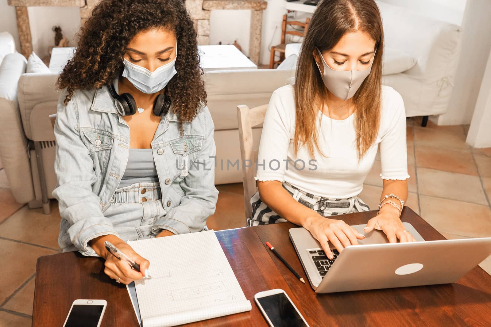 Two young mixed race beautiful women working from home sitting on a wooden table in elegant living room wearing Coronavirus safe protection mask - College female students using laptop to study online