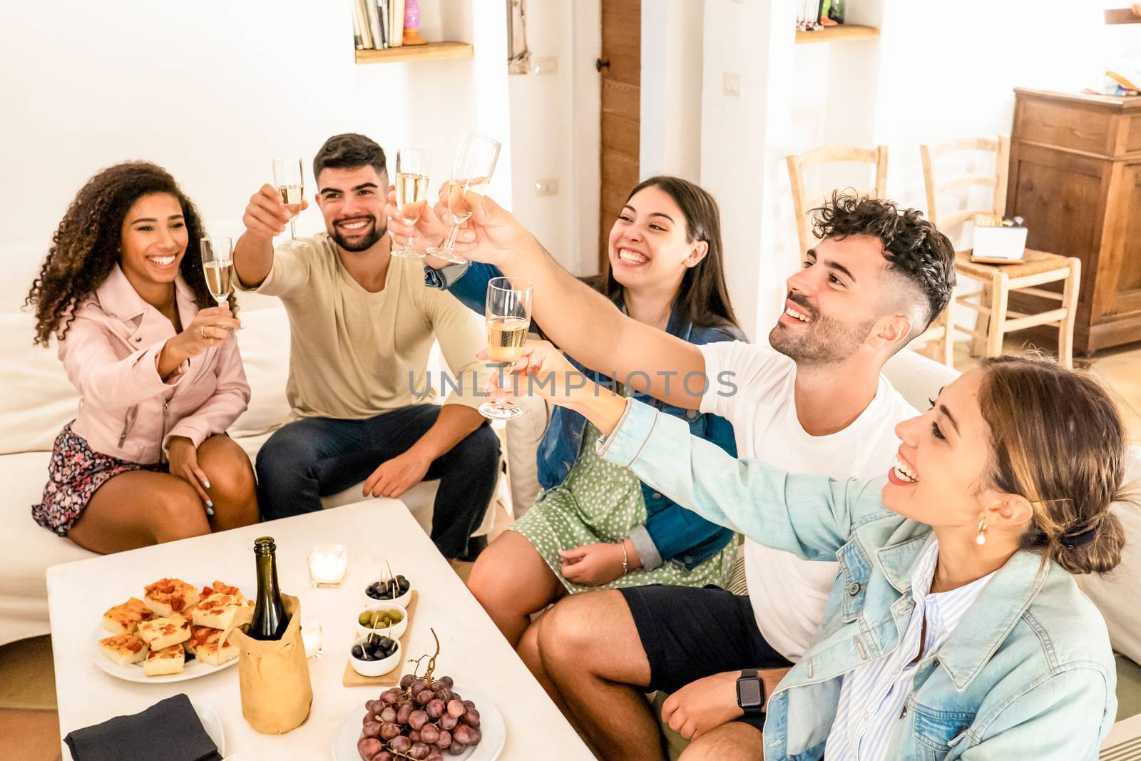 Group of mixed race friends toasting with champagne celebrating at home - Young multiracial people sitting on the sofa clinking glasses of white wine with table laden with snacks - Focus on glasses by robbyfontanesi