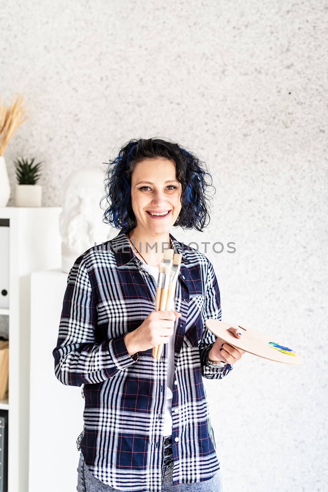 Smiling woman artist in her studio holding art palette and paintbrushes by Desperada