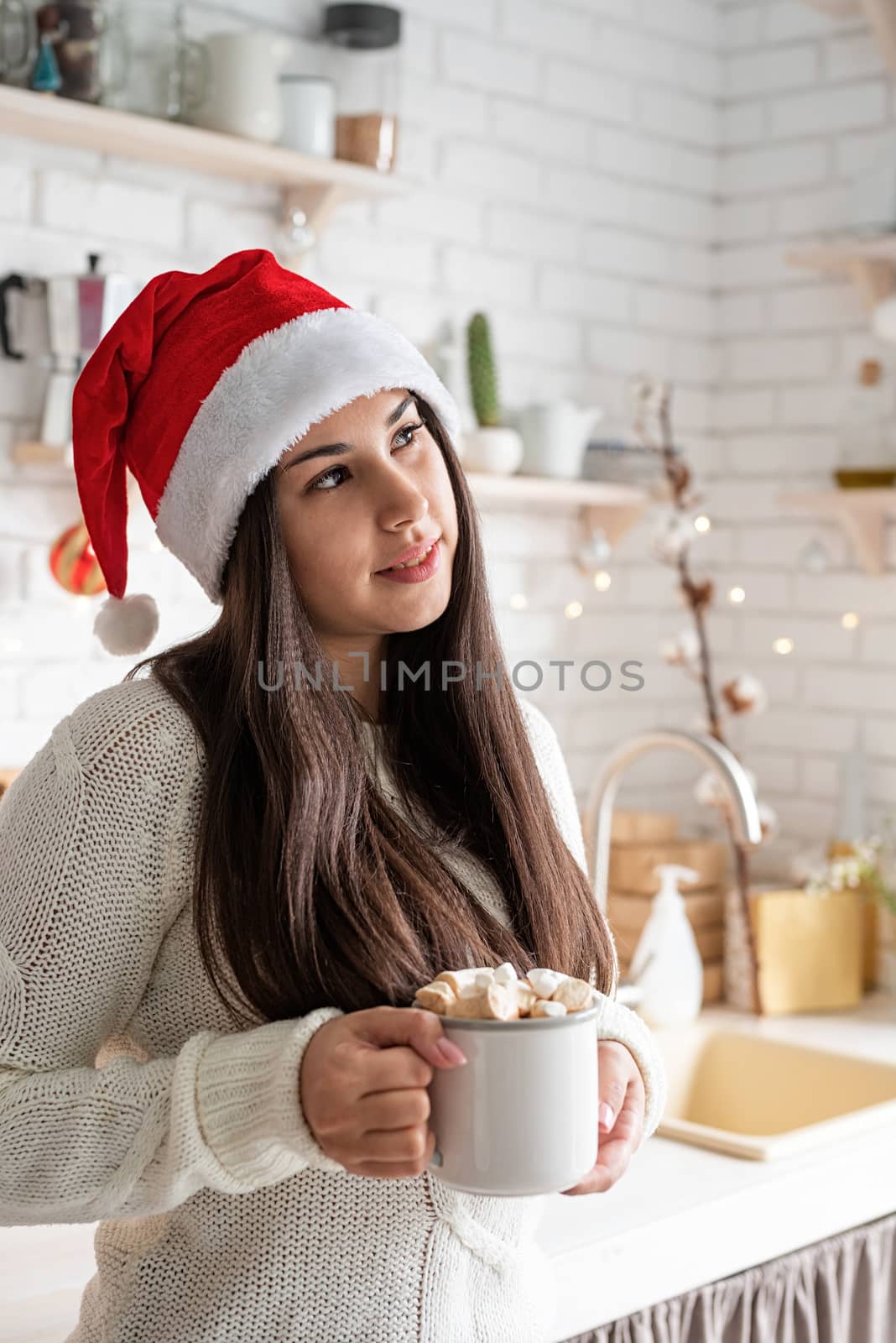 Christmas and New Year. Brunette woman in santa hat holding a cup of marshmallow cocoa in the kitchen