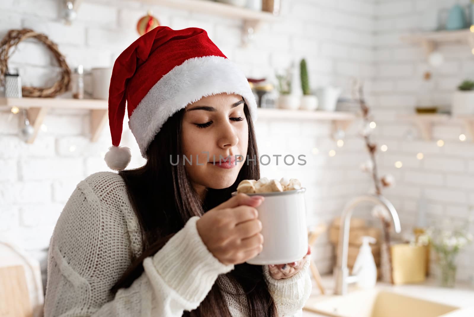 Brunette woman in santa hat holding a cup of marshmallow cocoa in the kitchen by Desperada