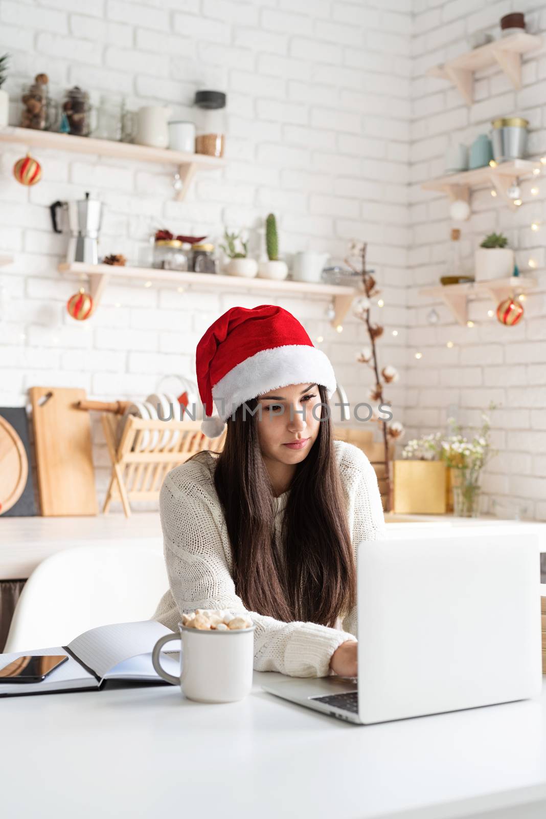 Merry Christmas and Happy New Year. Young brunette woman in santa hat chatting with friends using her laptop at the kitchen