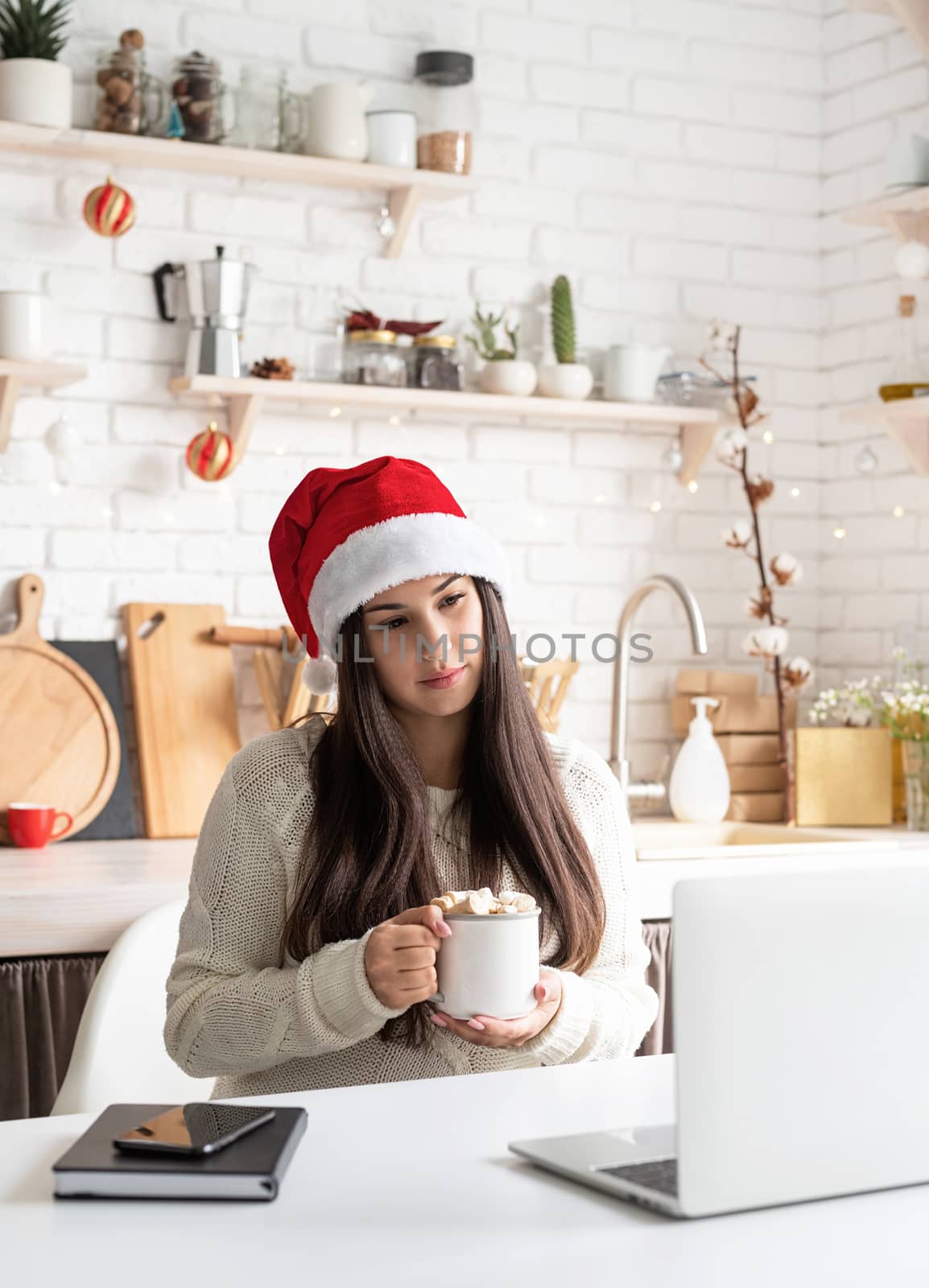 Young brunette woman in santa hat chatting with friends using her laptop at the kitchen by Desperada