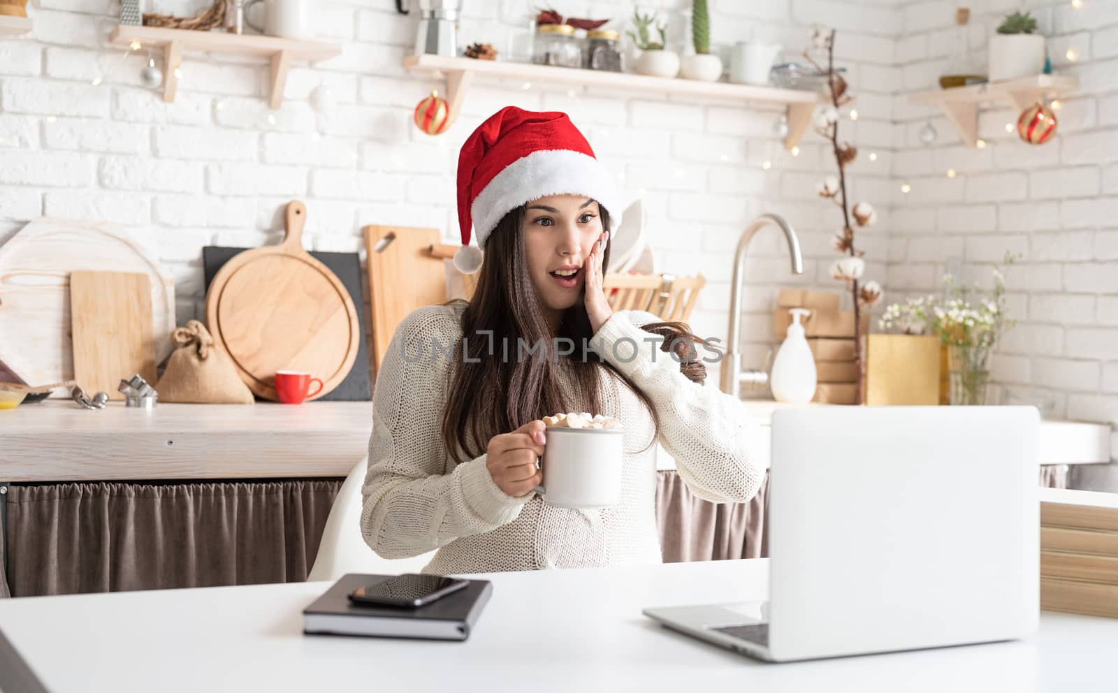 Merry Christmas and Happy New Year. Young surprised brunette woman in santa hat chatting with friends using her laptop at the kitchen