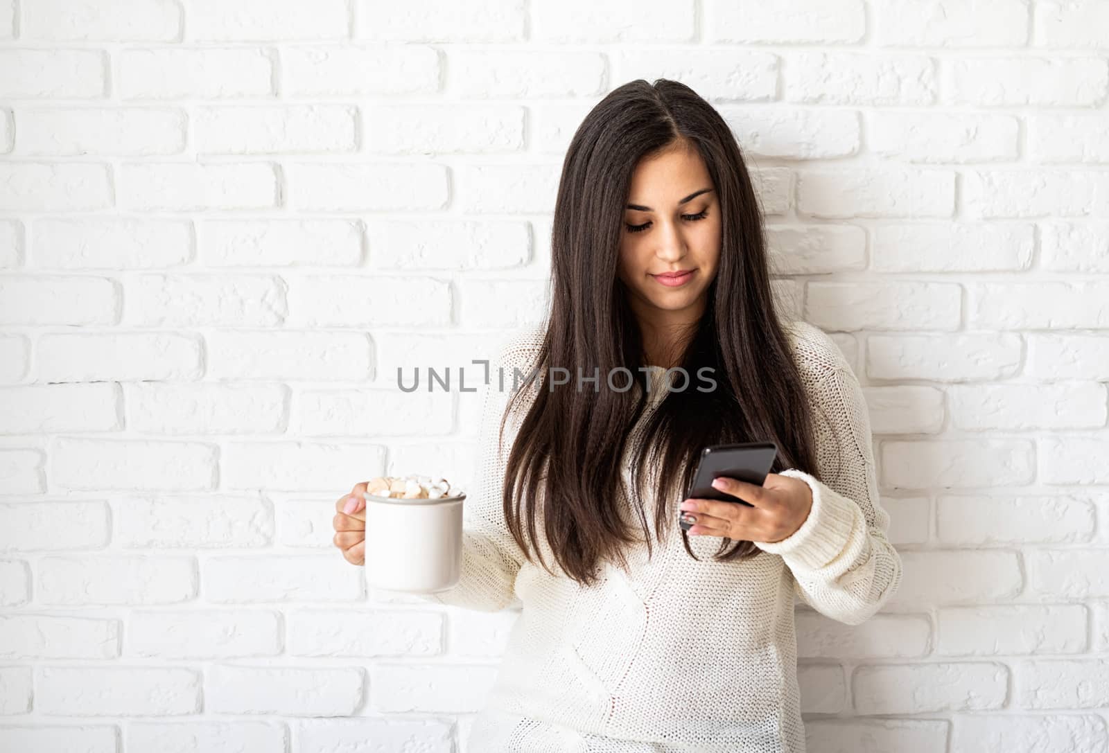 Young brunette woman holding a cup of marshmallow cocoa and using her mobile phone on white brick wall background by Desperada