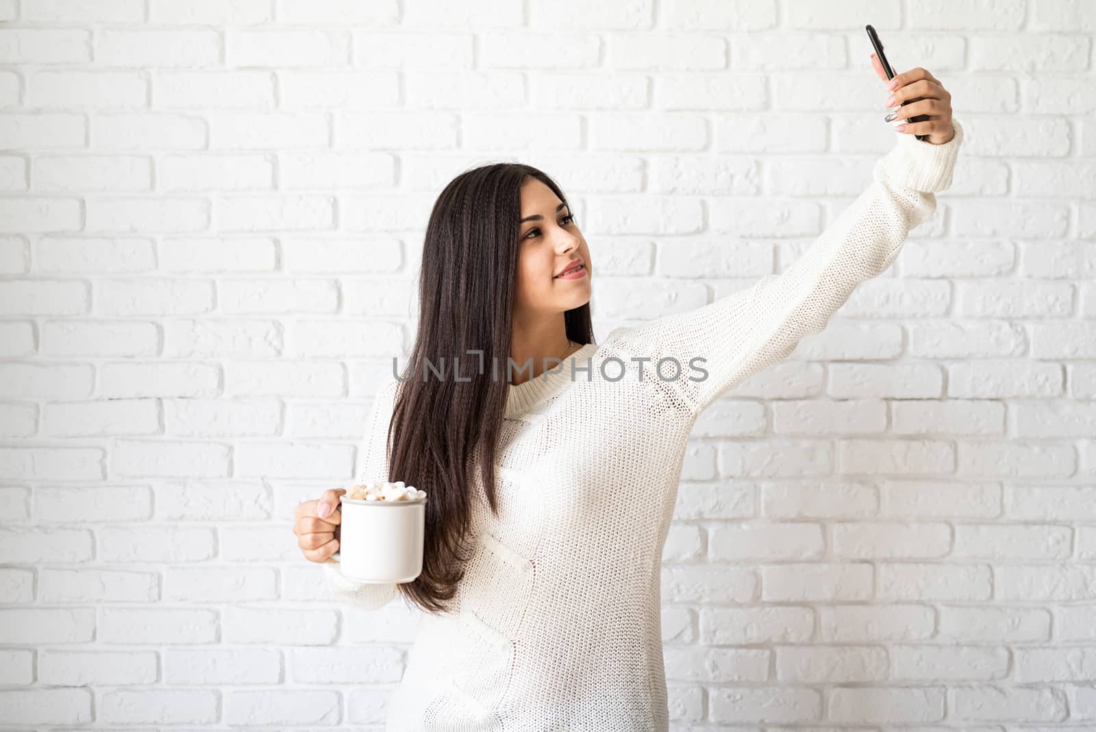Christmas and New Year. Young brunette woman holding a cup of marshmallow cocoa and taking selfie on white brick wall background