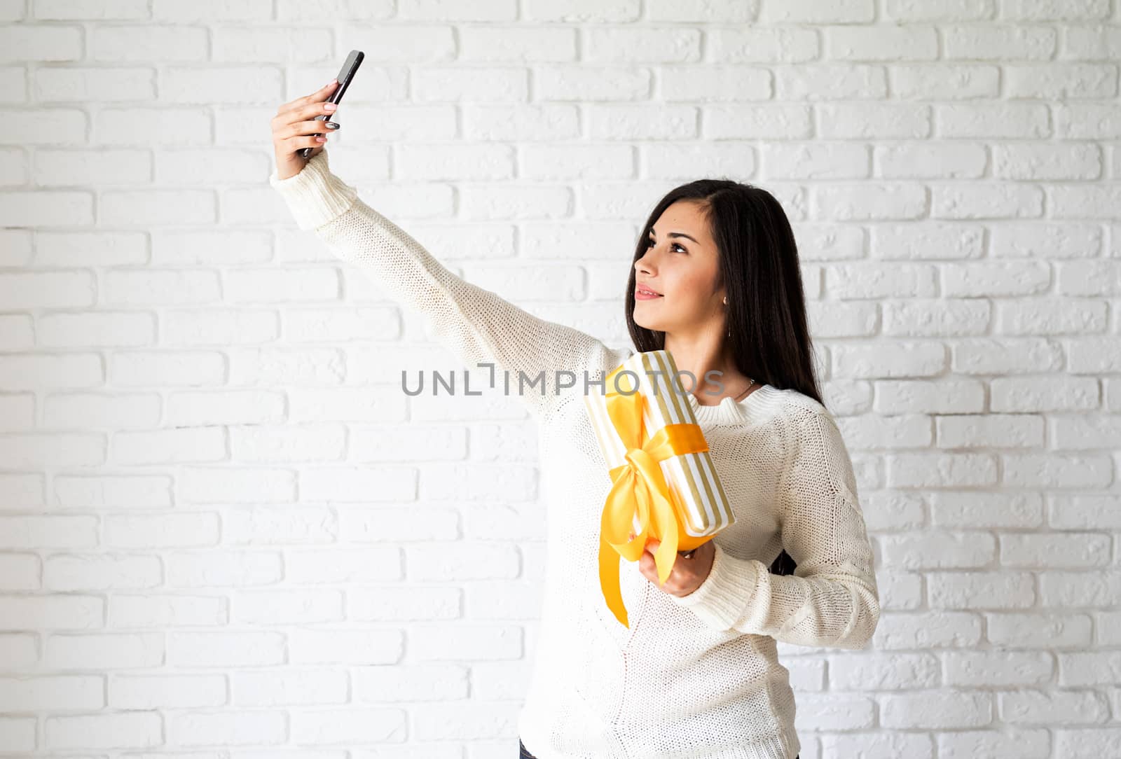 Young brunette woman holding a gift box and taking selfie on white brick wall background by Desperada