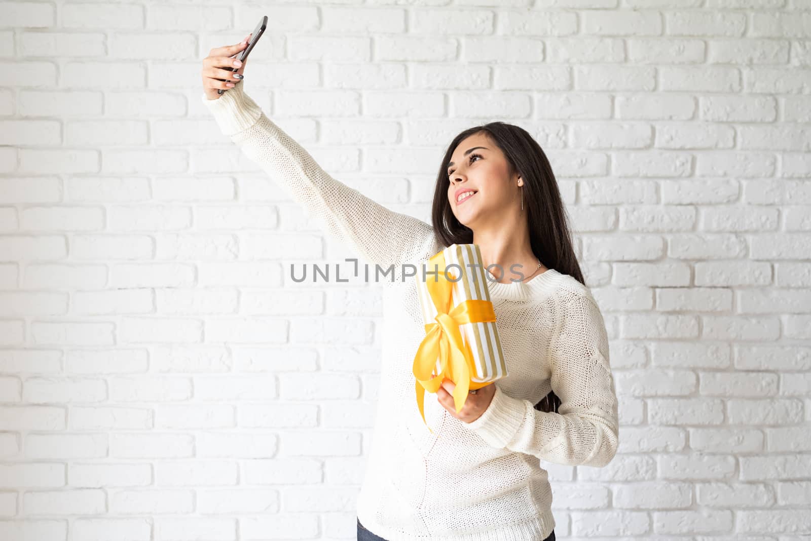 Christmas and New Year. Young brunette woman holding a gift box and taking selfie on white brick wall background