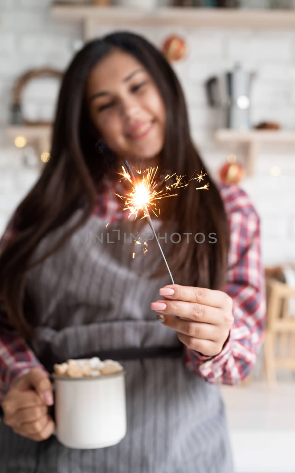 Young smiling woman with a cup of marshmallow cocoa and a sparkler celebrating Christmas in the kitchen by Desperada