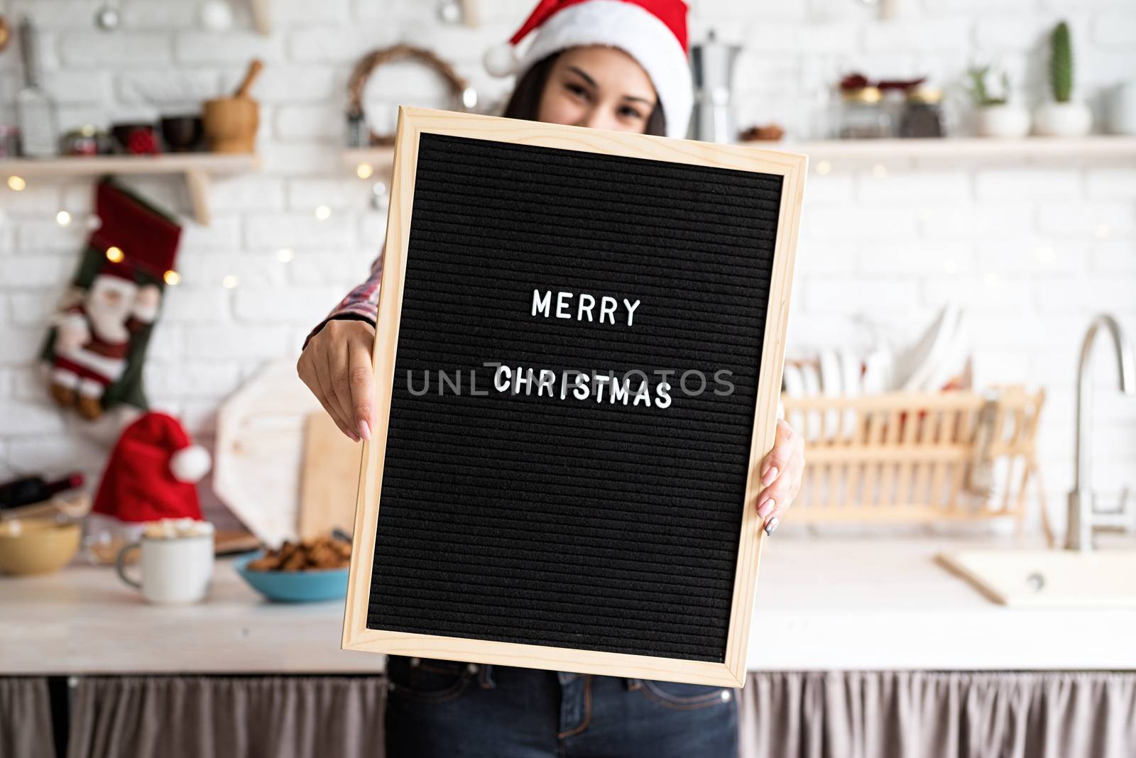 Merry Christmas and Happy New Year. Portrait of a happy beautiful latin woman in santa hat with black letter board with the words Merry Christmas on home kitchen background