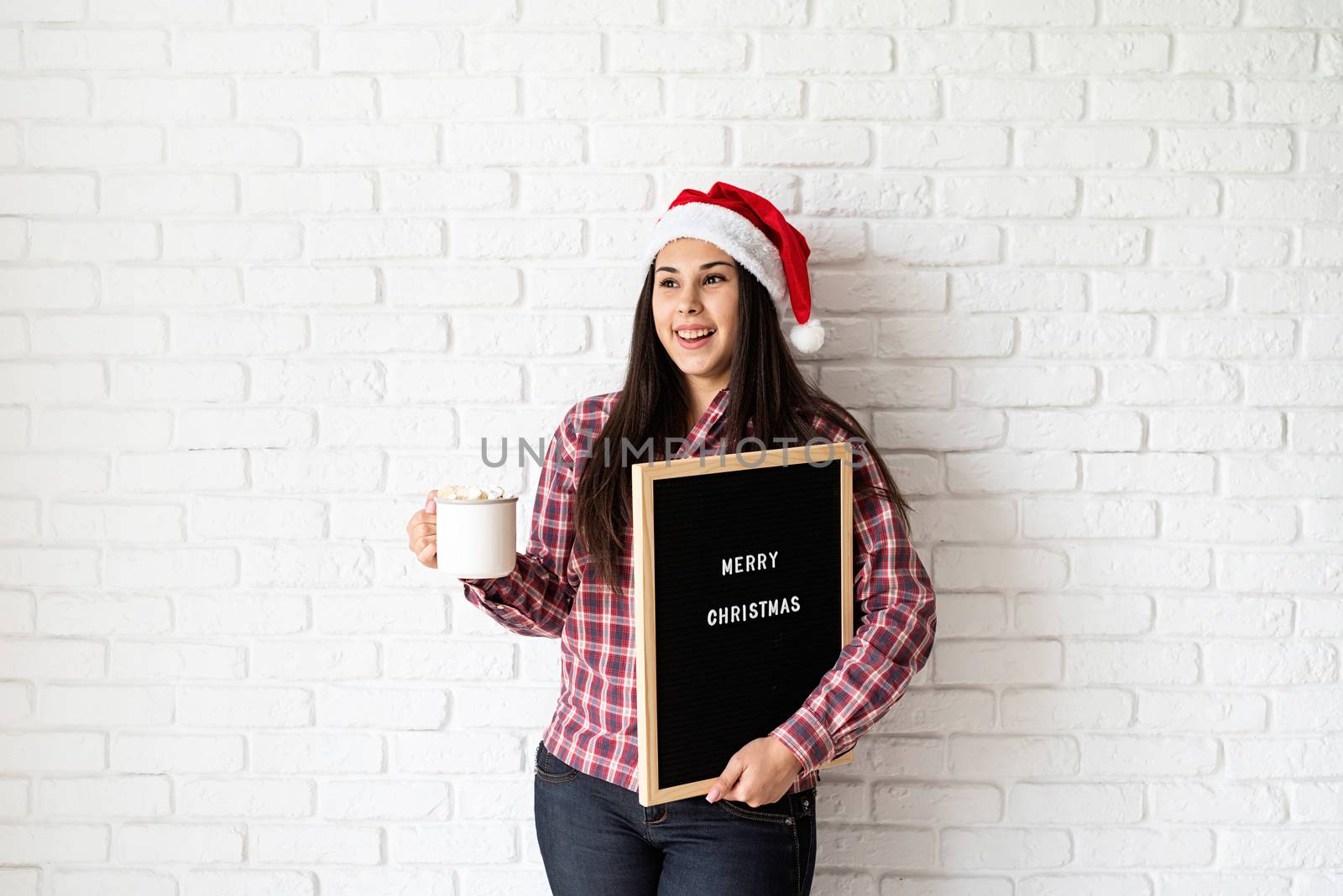 Portrait of a happy beautiful latin woman with letter board with the words Merry Christmas holding a cup of cocoa with marshmallows by Desperada