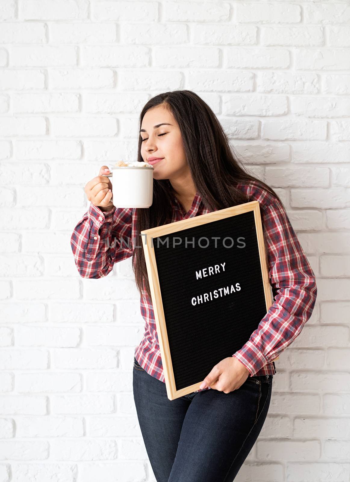 Portrait of a happy beautiful latin woman with letter board with the words Merry Christmas holding a cup of cocoa with marshmallows by Desperada