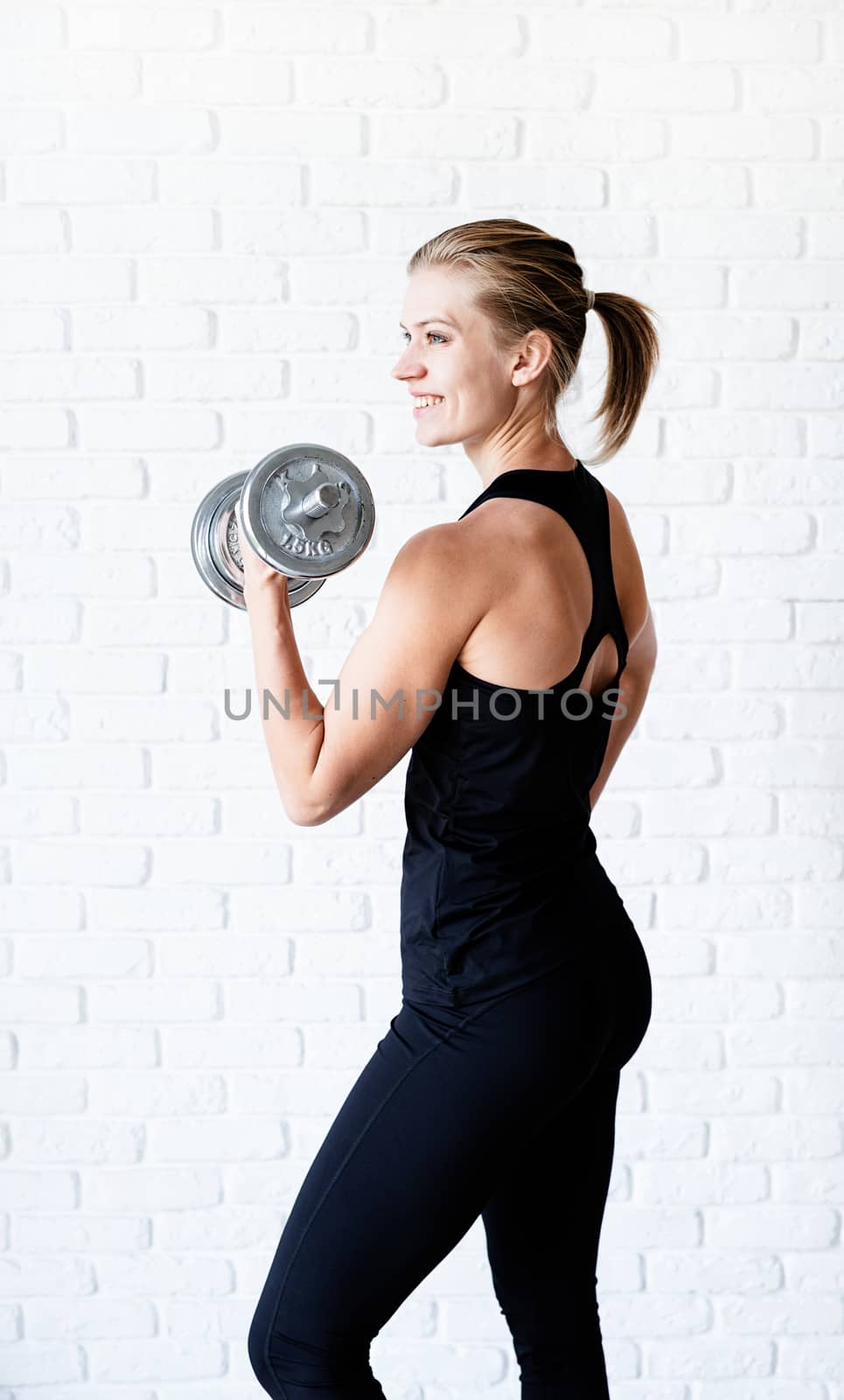 Portrait of athletic woman in black sportwear showing her muscles training with a dumbbell by Desperada