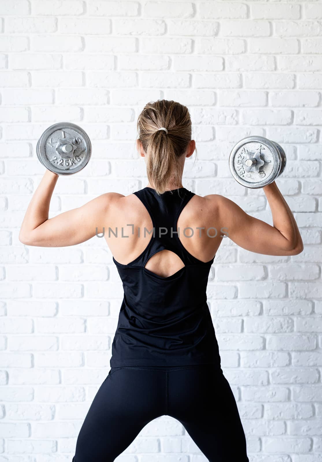 Rear view of an athletic woman in black sportwear showing her back and arms muscles training with a dumbbell by Desperada