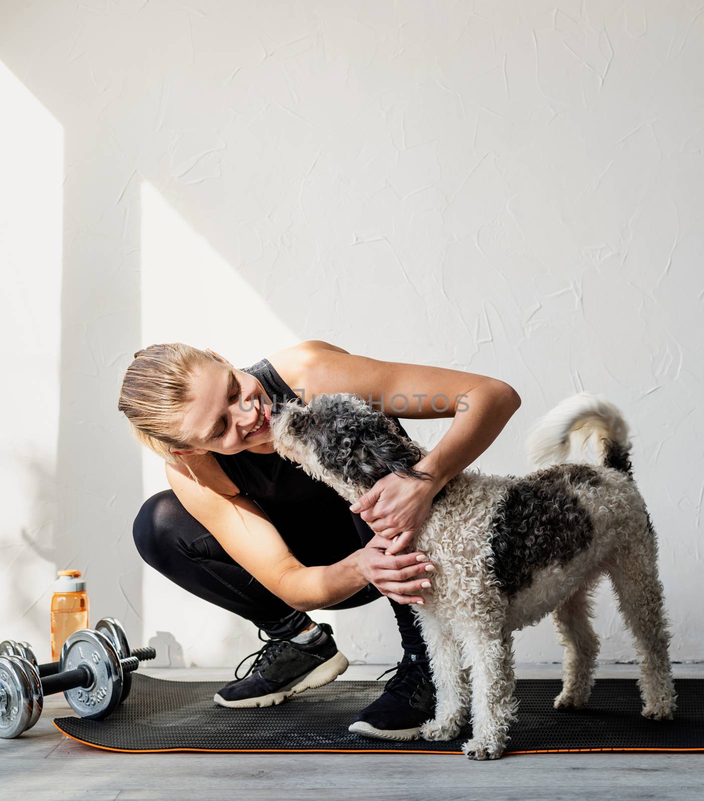 Happy young blond woman patting her dog at the home gym by Desperada