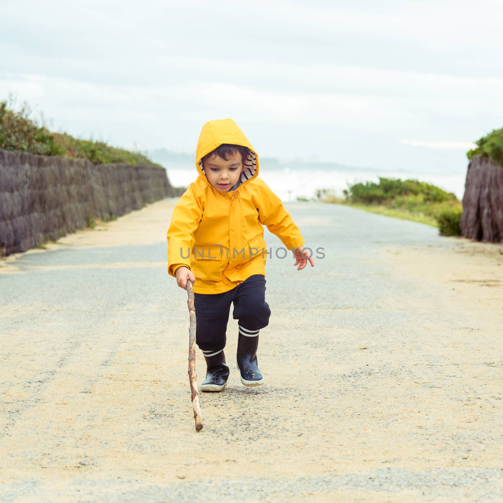 Little boy playing with wood stick, toddler wearing yellow raincoat and gumboots, coastal area