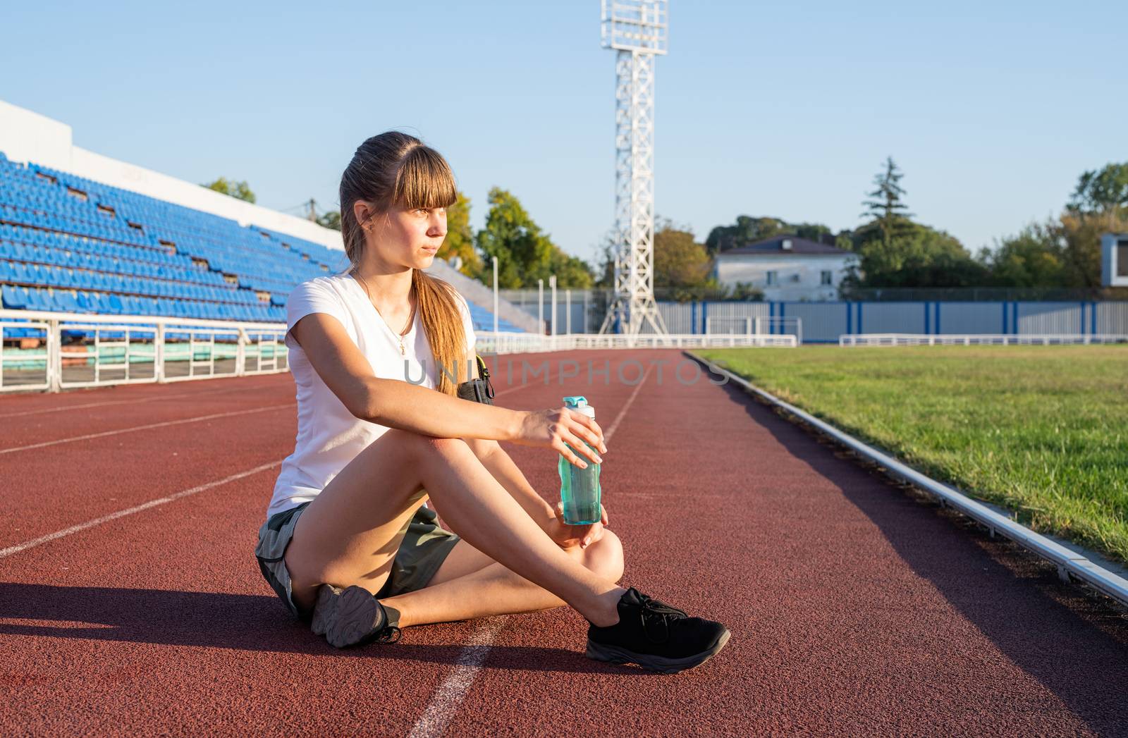 Sports and fitness. Teenager girl sitting on stadium track having rest drinking water