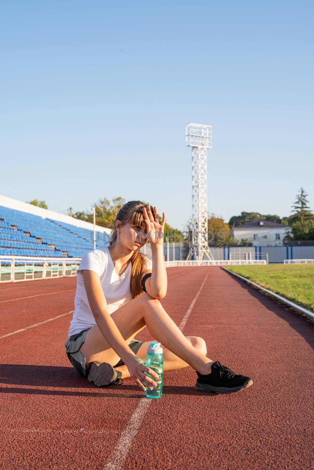 Sports and fitness. Teenager girl sitting on stadium track having rest drinking water
