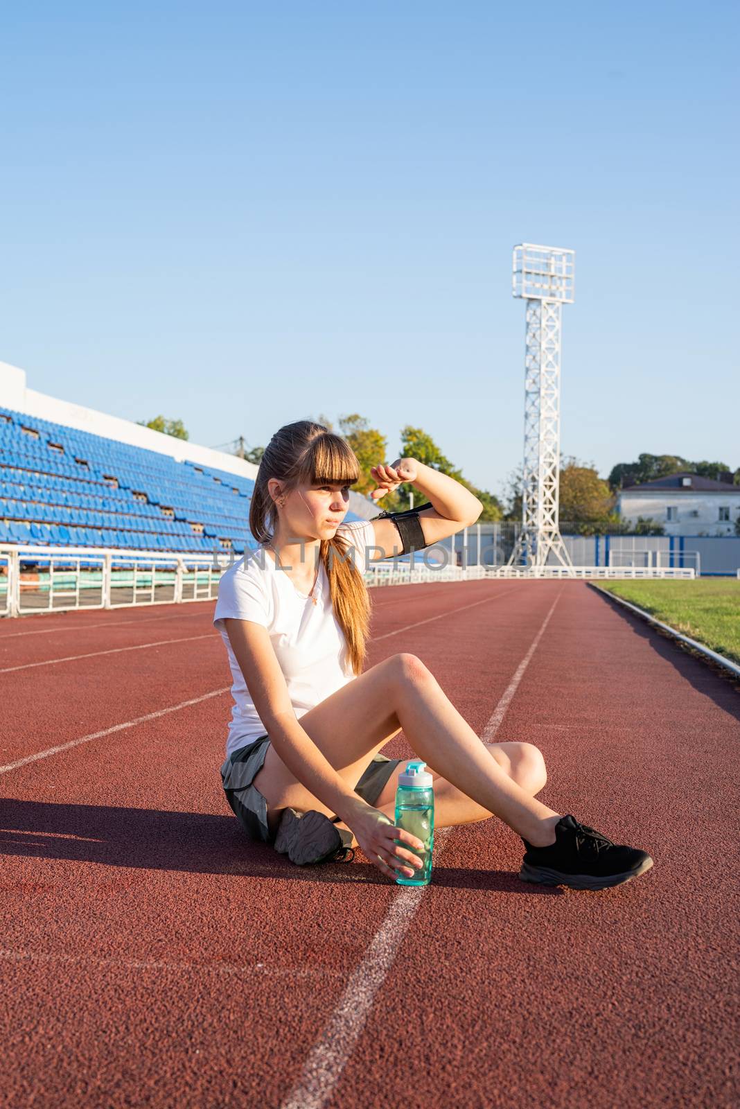 Teenager girl sitting on stadium track having rest drinking water by Desperada