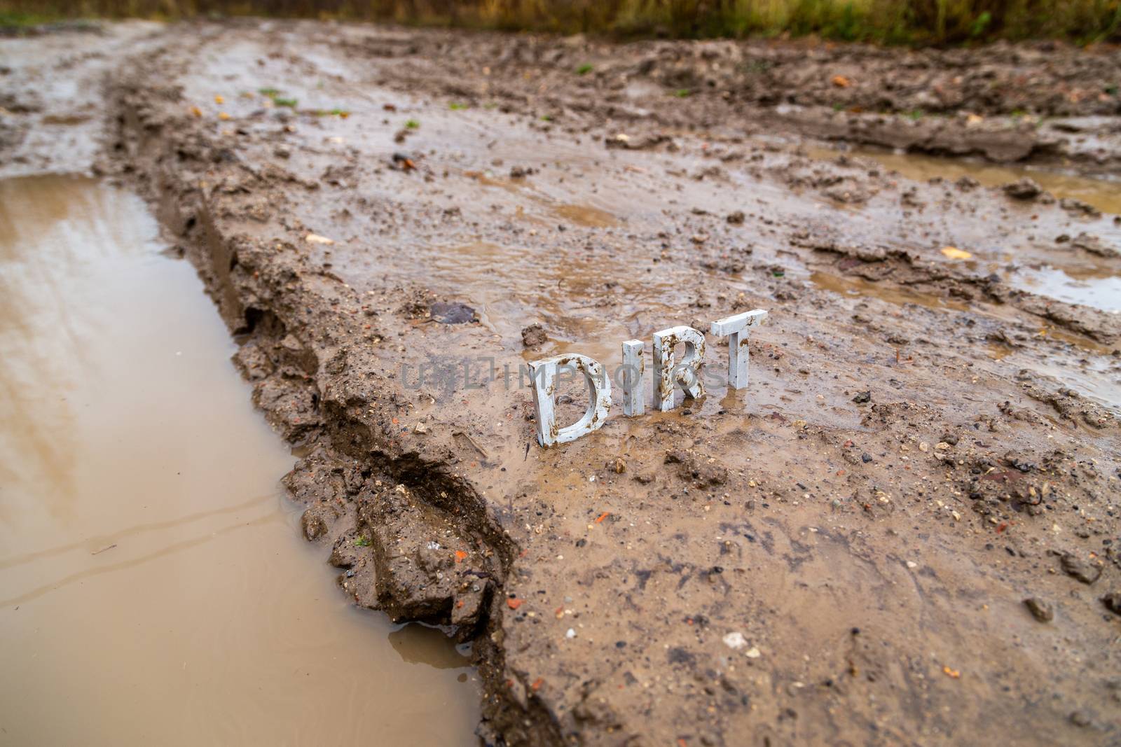 the word dirt composed of silver metal letters on wet clay surface with selective focus, background blur and linear perspective in cloudy autumn day light