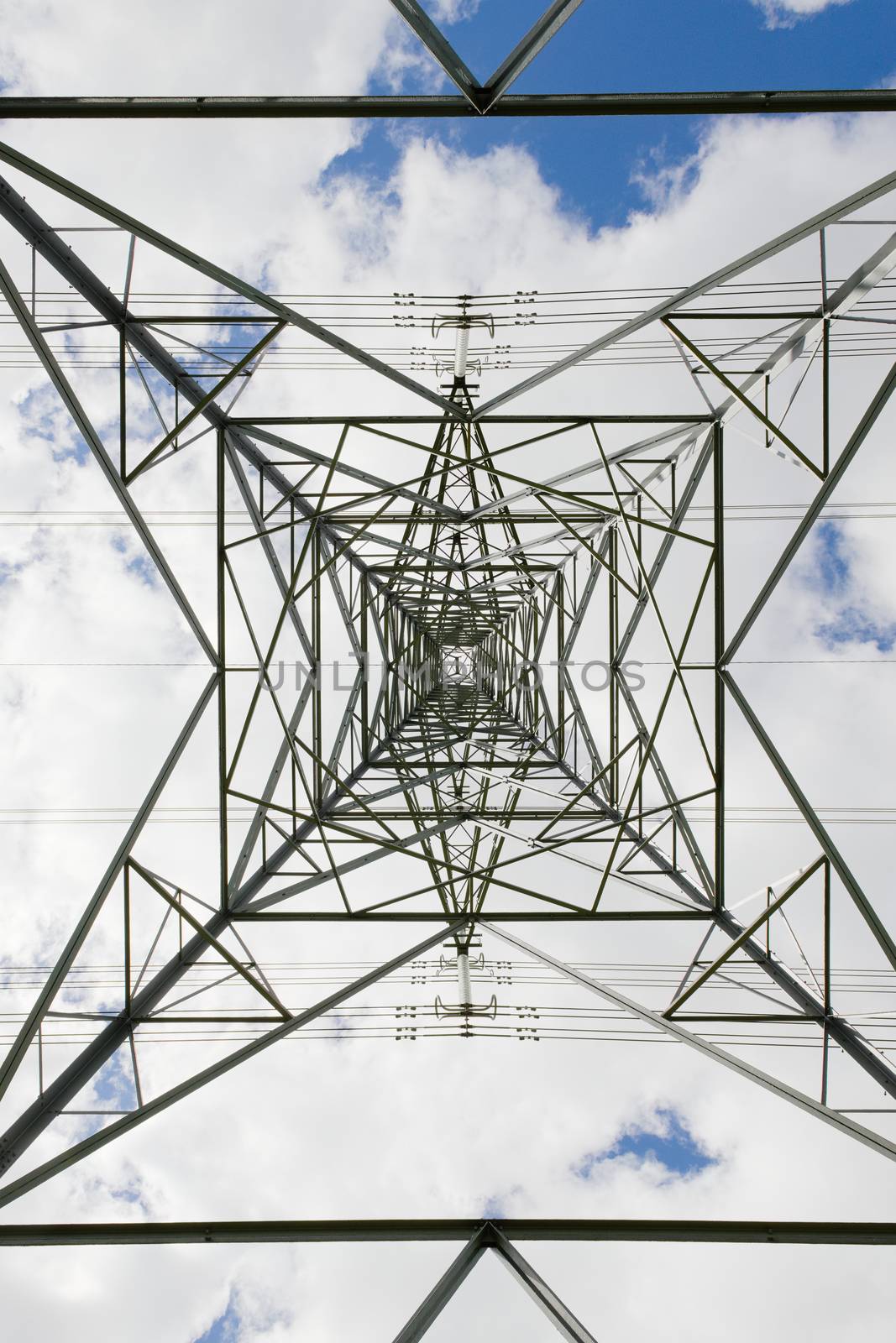 Converging symmetrical patterns created by the metal sheet lattice work that makes up an electrical transmission pylon tower. Blue cloudy sky behind