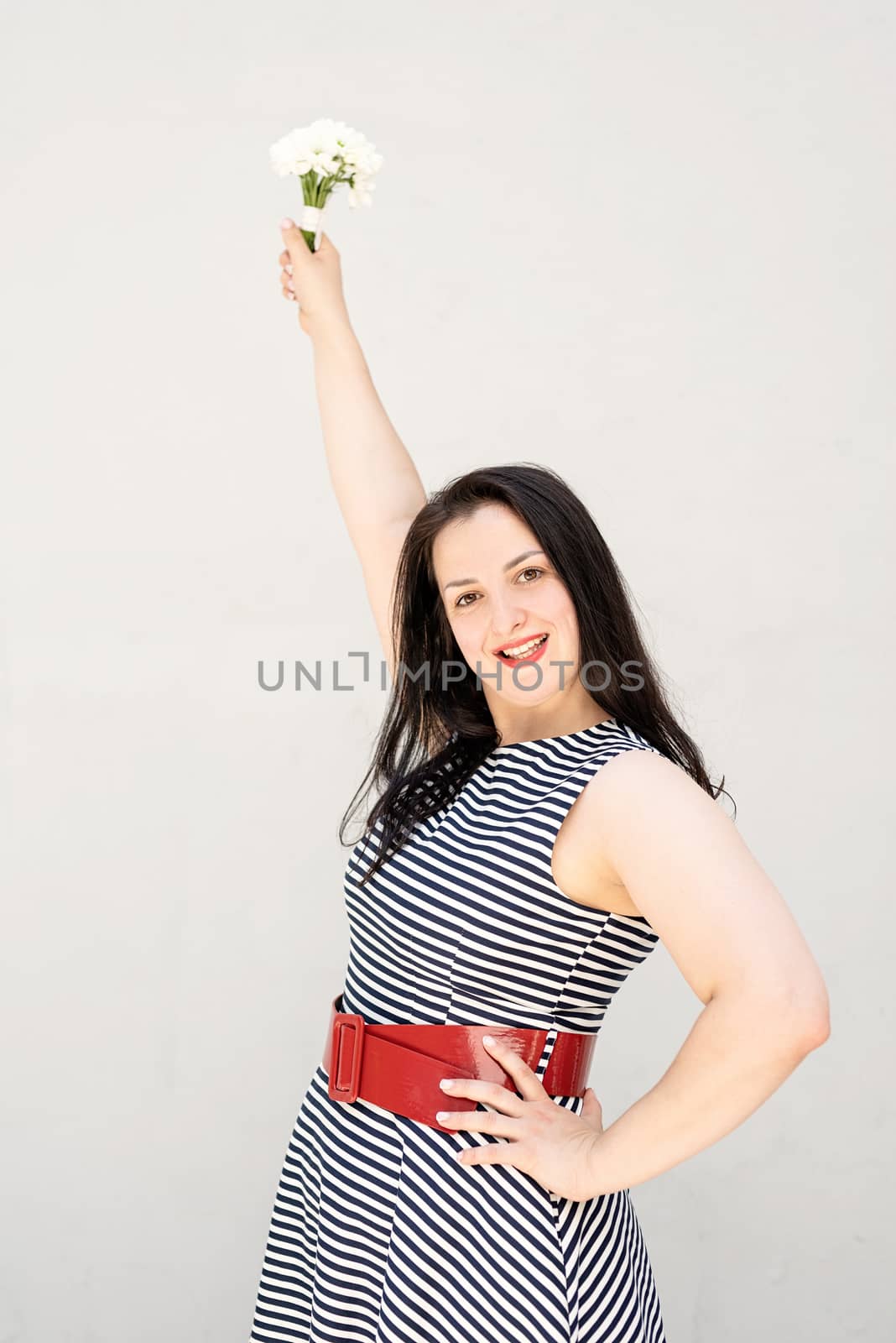 Happy young woman holding a bouquet of flowers in a raised hand on a gray solid background by Desperada
