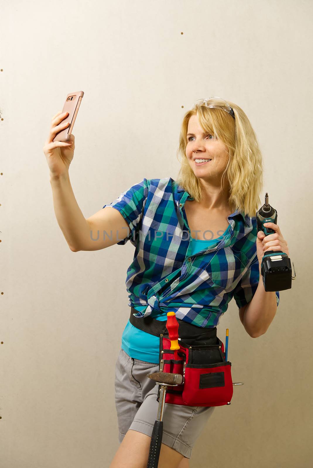 Blond woman wearing a DIY tool belt full of a variety of tools on a unpainted plasterboard wall background. Construction woman concept. by PhotoTime