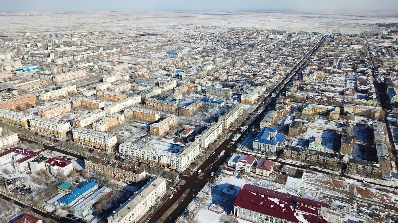 A small town on the shore of lake Balkhash. Top view of houses, cultural buildings, snowy streets, gray roads with cars and a green lake. The lake is partially covered with ice. Soviet city.