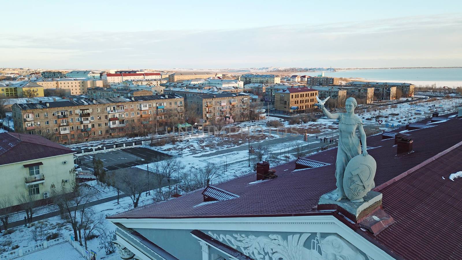 Statue of a woman with a shield on the roof of the Palace. Red tiles on the roof. View of the lake, houses and sunset. The small town of Balkhash in winter.
