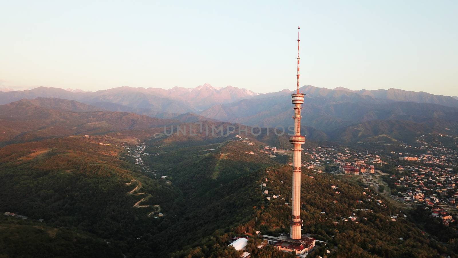 Kok Tobe big TV tower on the green hills of Almaty. View of the mountains, sky, green hills, houses and road. People are walking around the square and alleys below. Houses are visible in the distance