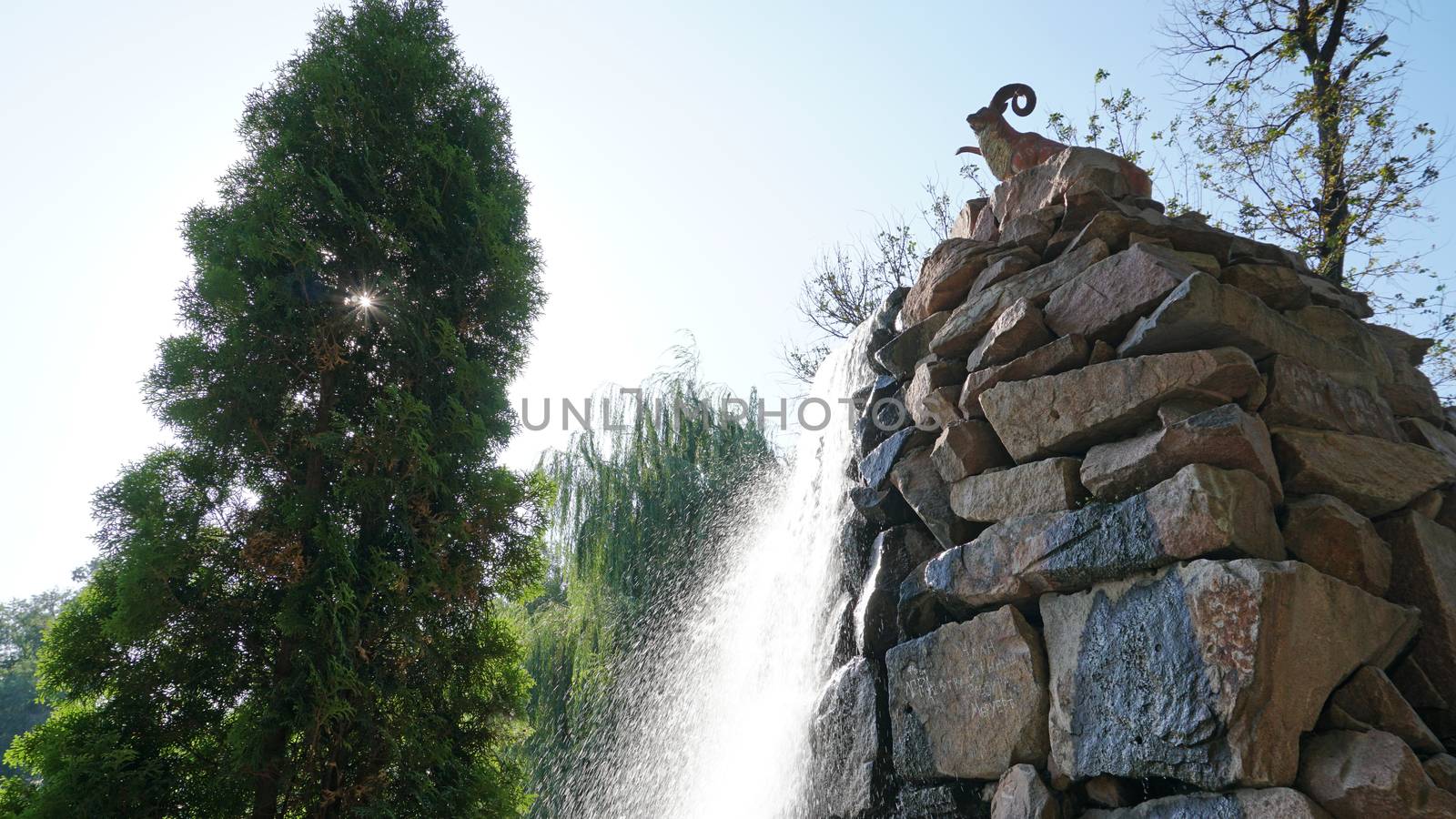 Artificial waterfall with a statue in the city Park of Almaty. Mountain sheep stands on the high stones of the waterfall, water flows into the bowl. Green trees and sky all around.