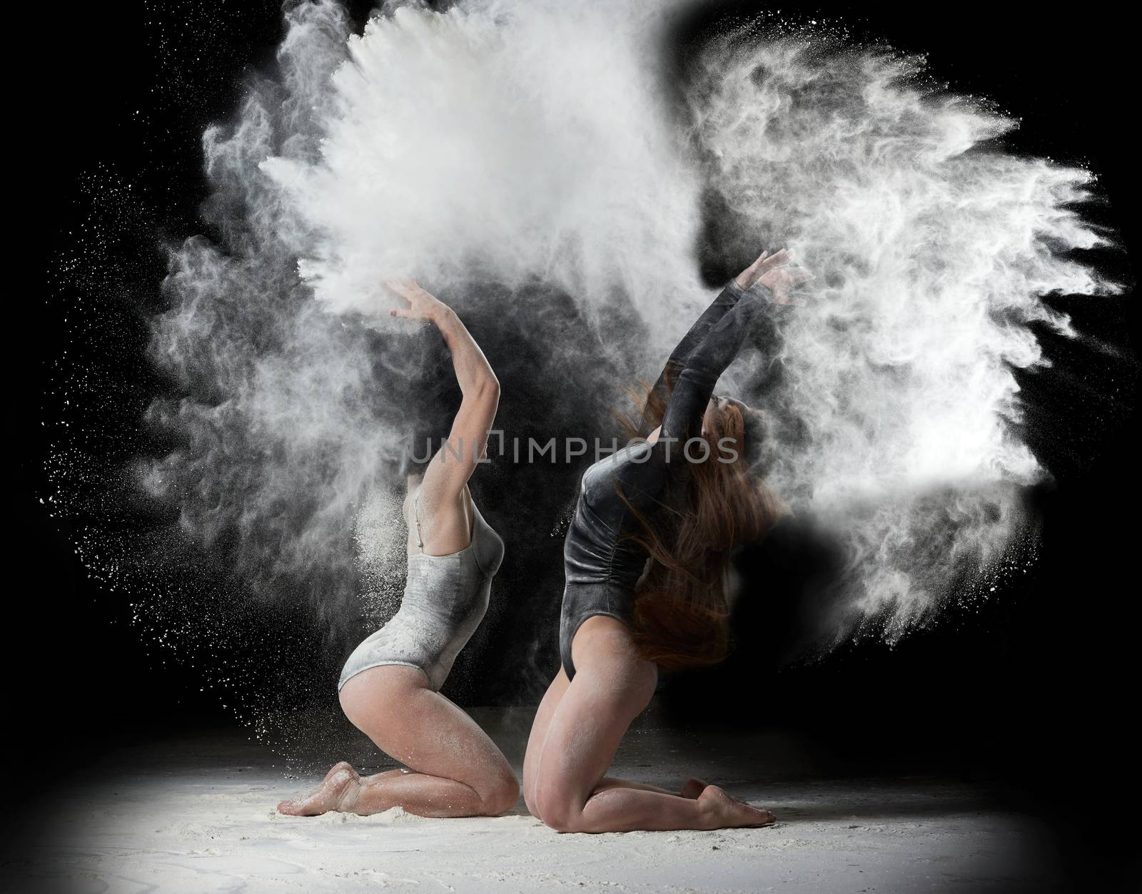 two beautiful young caucasian women in black bodysuits with a sports figure are dancing in a white cloud of flour on a black background, explosion and expresse in motion