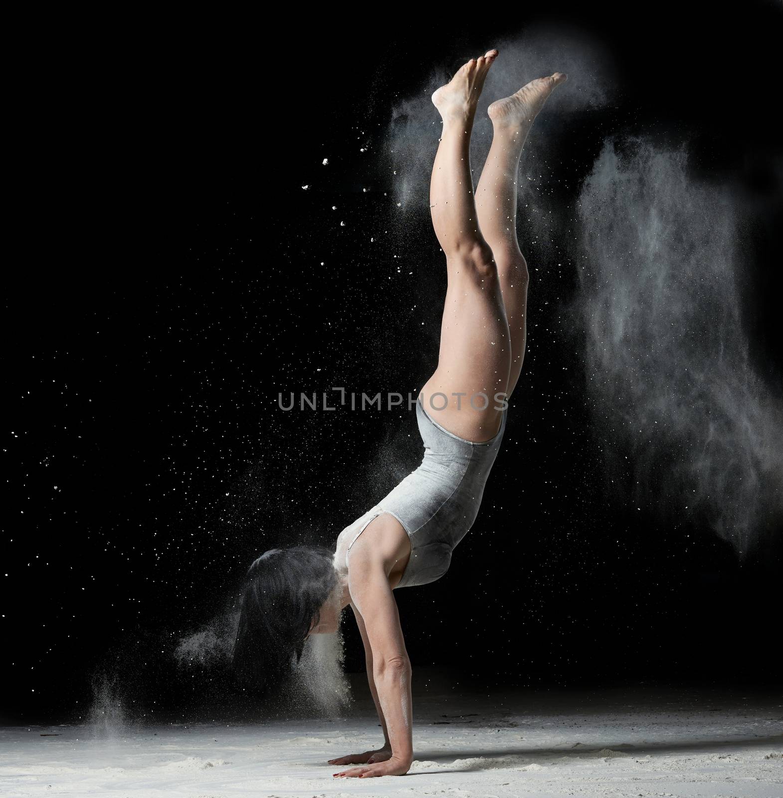 young woman with an athletic figure dressed in a black bodysuit stands upside down on her hands, black background with white flying flour, acrobatic exercises