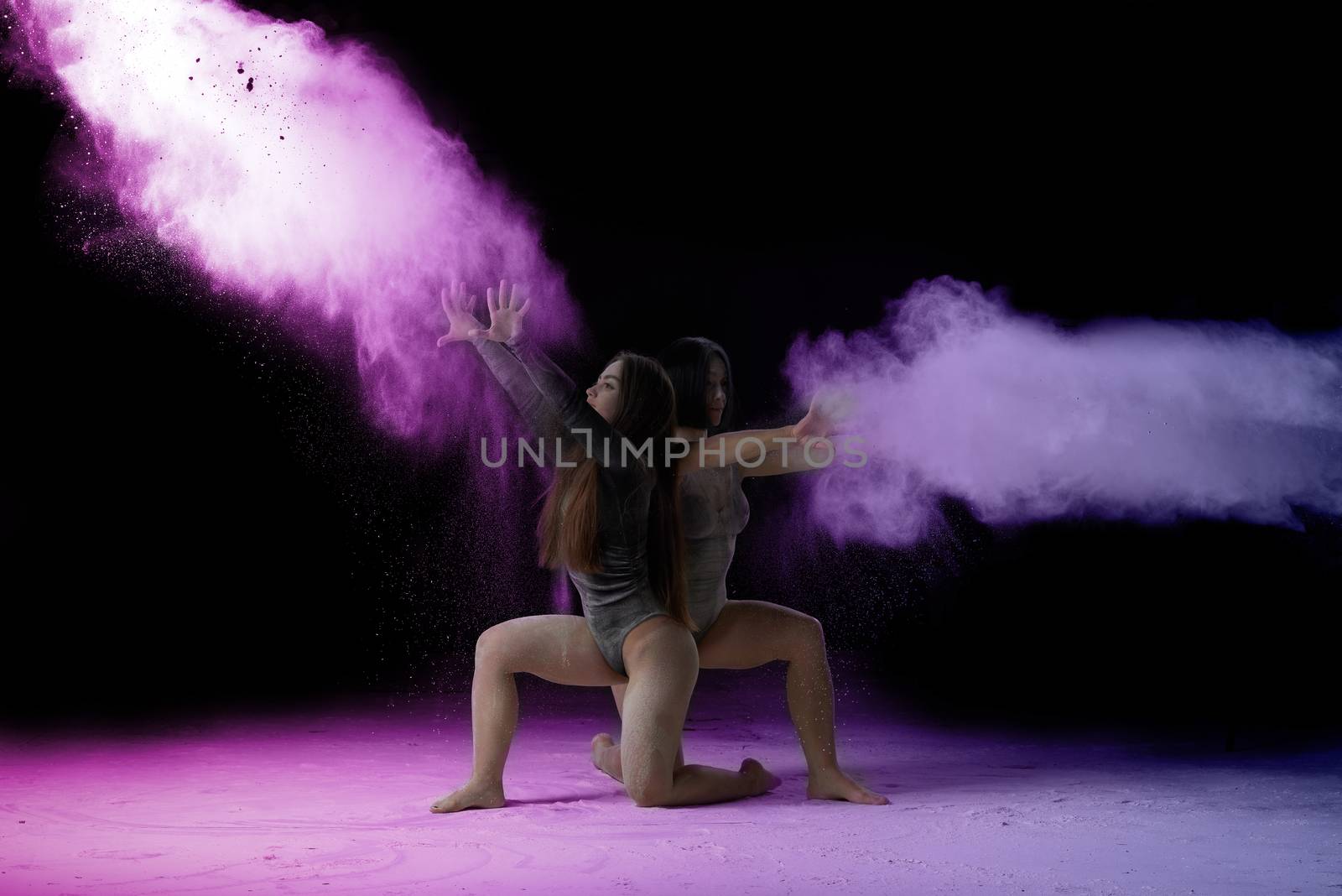 two beautiful young caucasian women in black bodysuits with a sports figure are dancing in a purple-blue cloud of flour on a black background, explosion and expresse in motion