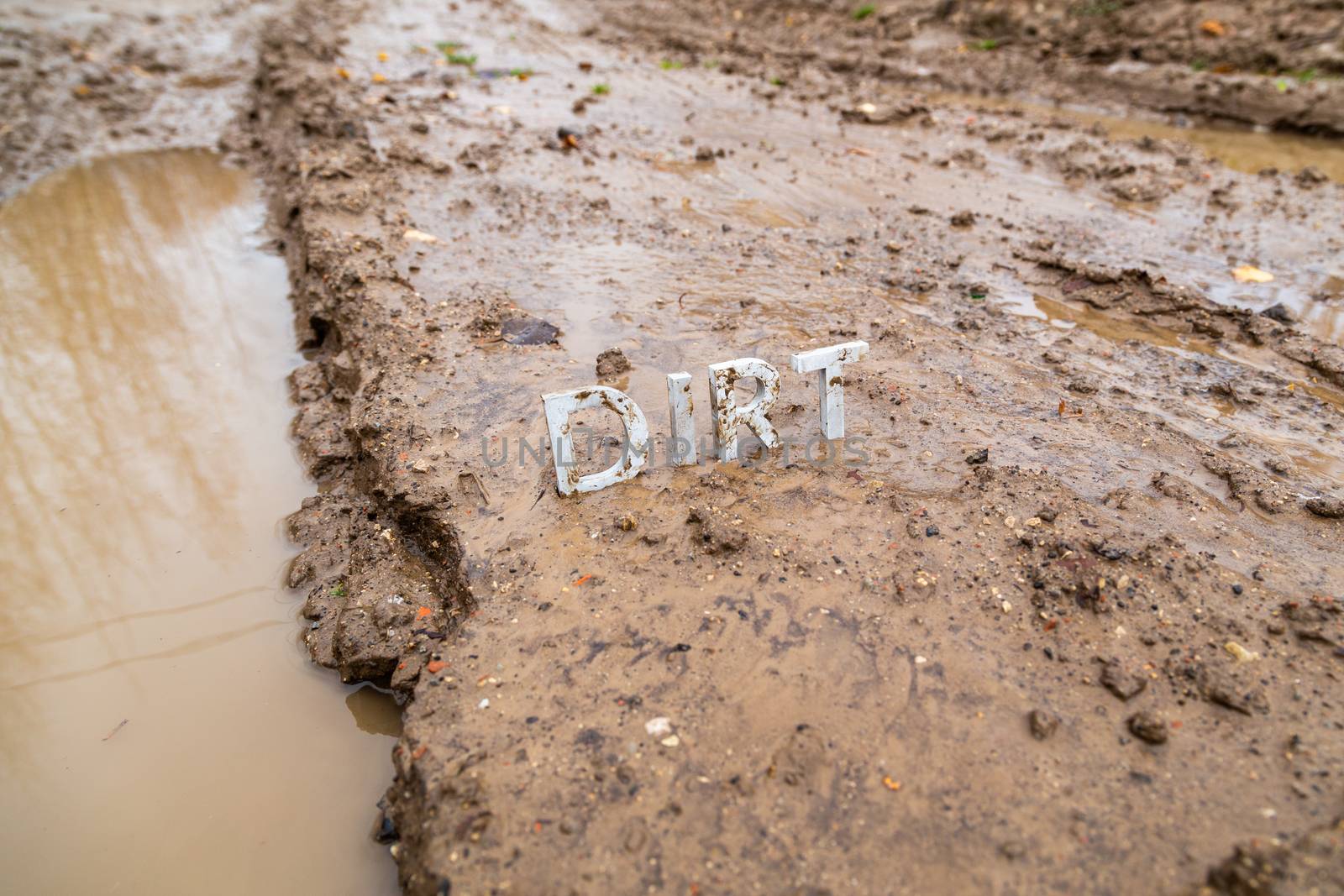 the word dirt composed of silver metal letters on wet clay surface with selective focus, background blur and linear perspective in cloudy autumn day light