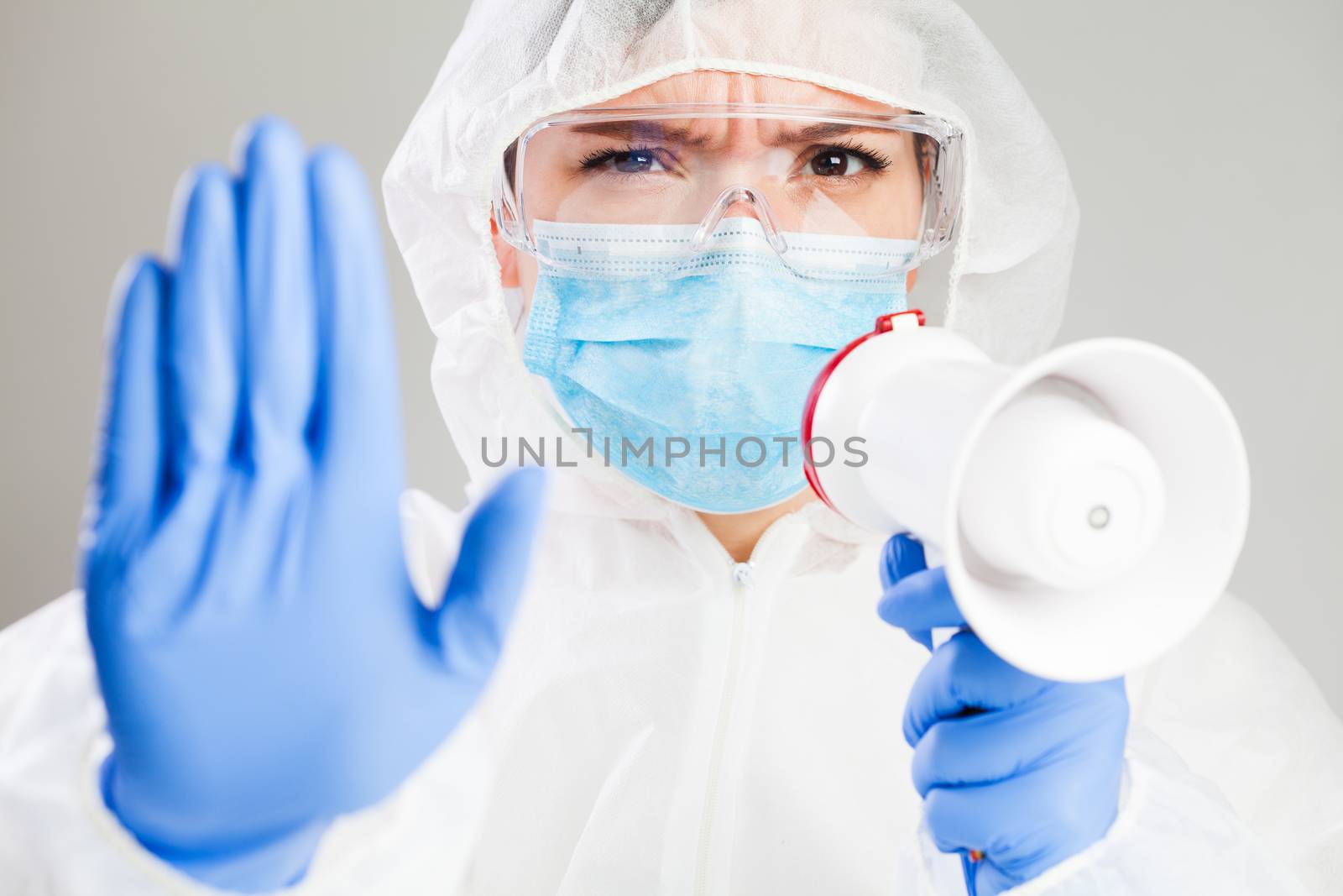 Doctor or medic in white protective suit, safety goggles, mask, gloves, holding a megaphone, shouting commands, raised arm with palm gesturing STOP, Coronavirus COVID-19 virus disease pandemic crisis