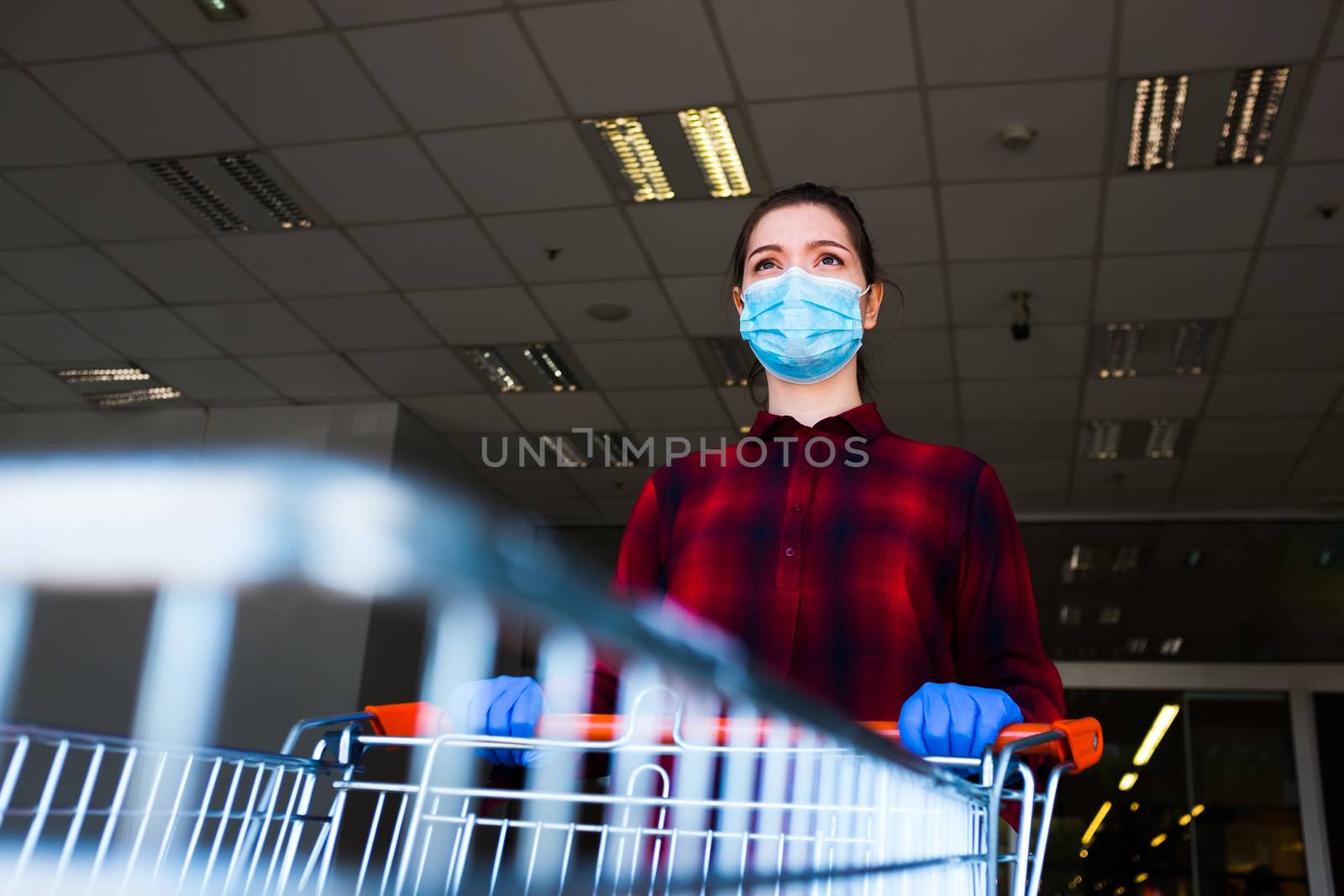 Worried and anxious caucasian woman wearing protective surgical face mask and latex gloves, pushing shopping cart exiting supermarket, global Coronavirus COVID-19 corona virus pandemic crisis outbreak