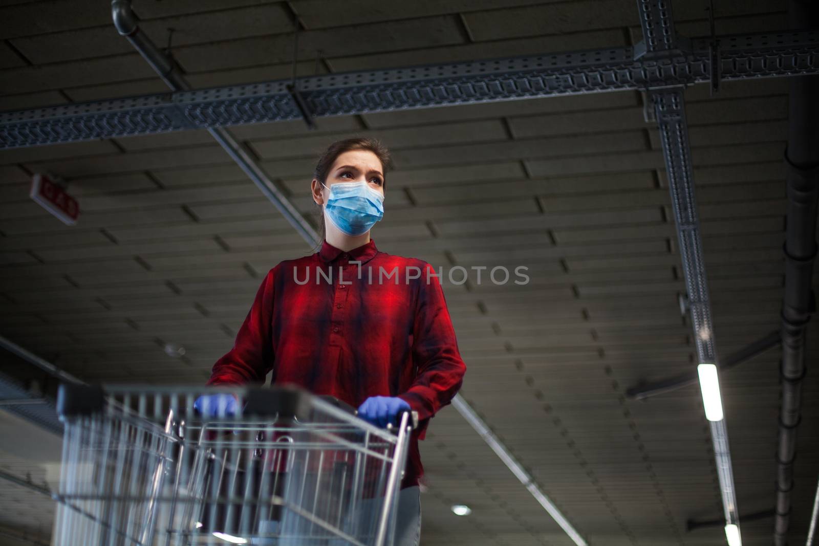 Caucasian woman pushing shopping cart in supermarket underground garage wearing gloves and mask, worried and anxious, panic buying and stocking supplies due to Coronavirus COVID-19 pandemic crisis