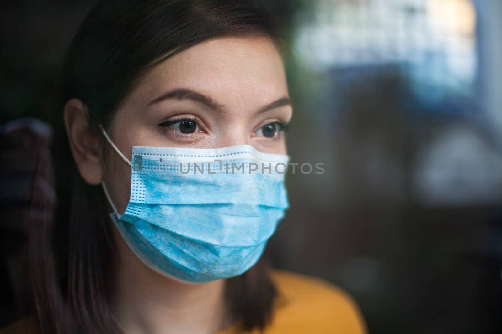 Anxious depressed and stressed young woman wearing medical face mask,in home isolation looking outside window, uncertain future due to global Coronavirus COVID-19 pandemic crisis, stay at home concept