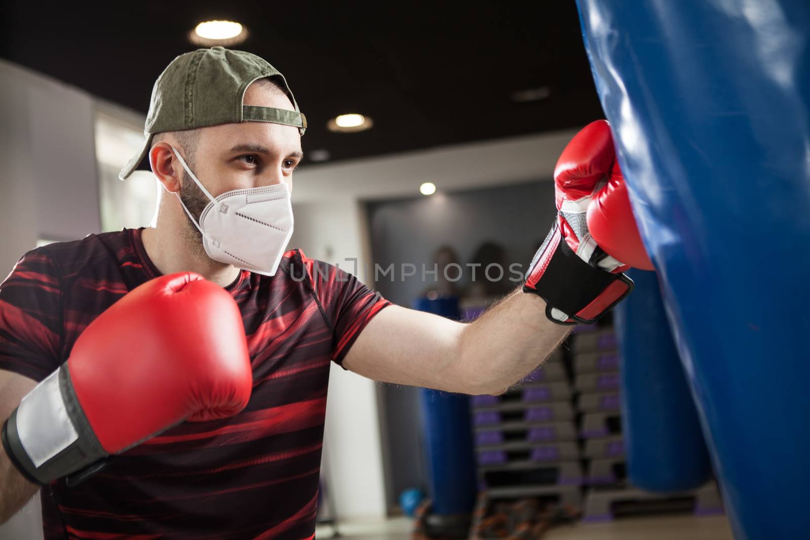 Young caucasian male wearing red boxing gloves & protective face mask,hitting blue sack in a reopen indoor gym,Coronavirus COVID-19 pandemic imposing social distancing rules and cautionary measures 
