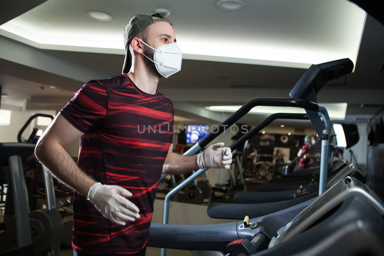 Young man running on treadmill in indoor gym,wearing protective face mask & rubber latex gloves,COVID-19 pandemic prevention of spread & transfer of Coronavirus,staying fit in lockdown concept in US