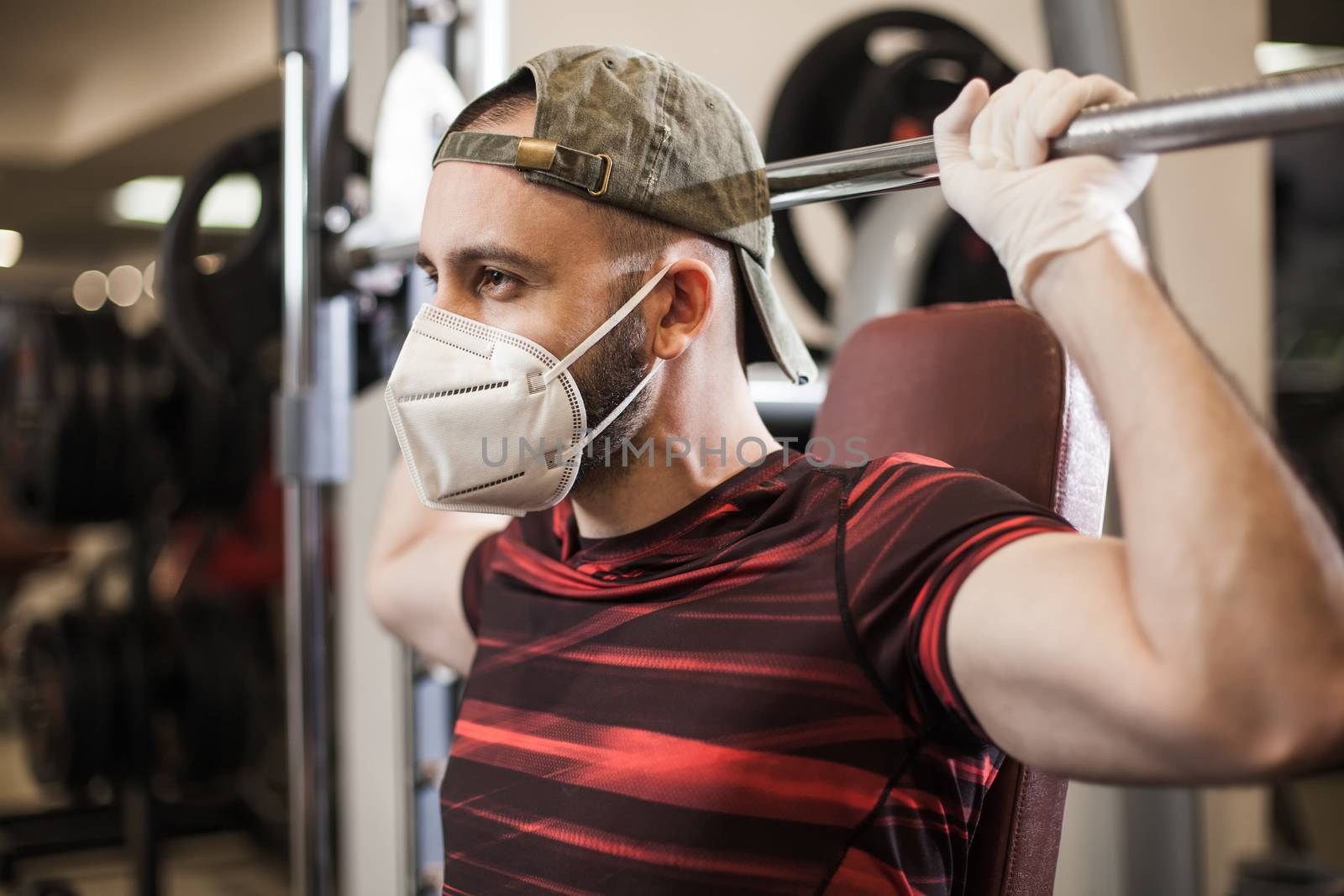 Young caucasian man lifting weights in US gym wearing protective rubber latex gloves & face mask,COVID-19 pandemic outbreak enforcing social distancing rule,prevention of Coronavirus spread & transfer