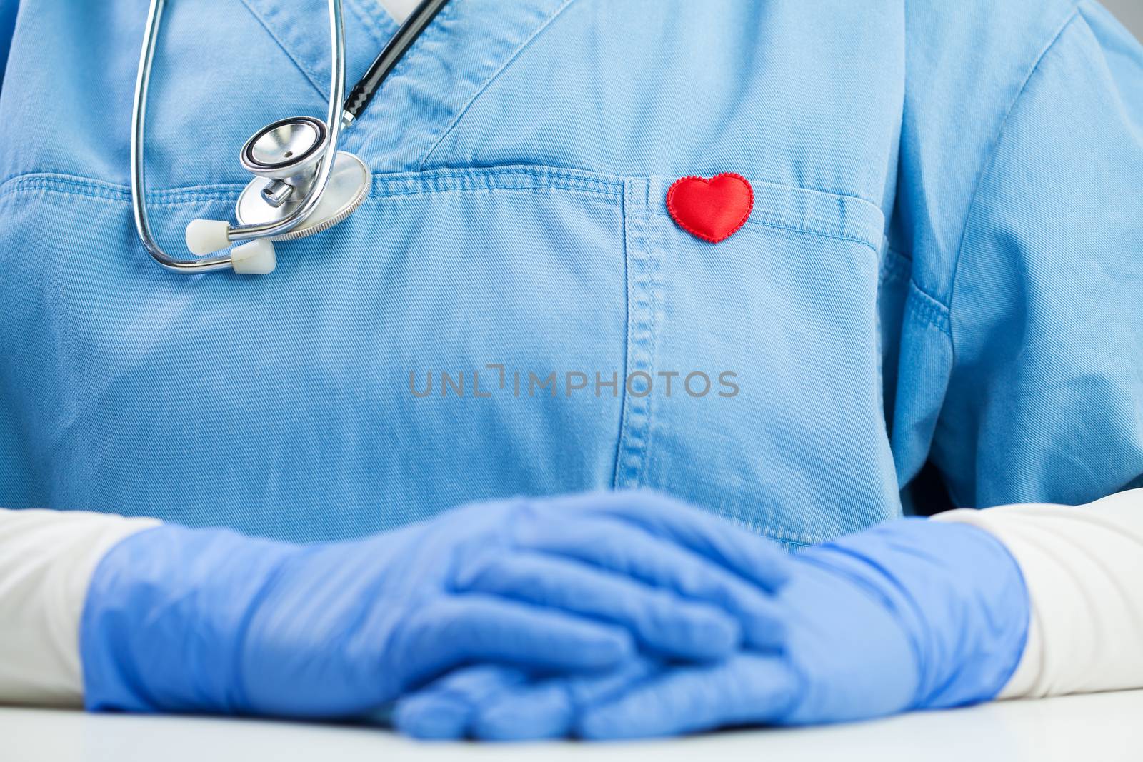 Doctor in blue uniform wearing a stethoscope and small red heart badge pin, detail closeup, GP or cardiologist in the office, sitting at the desk, Coronavirus COVID-19 global pandemic outbreak crisis 