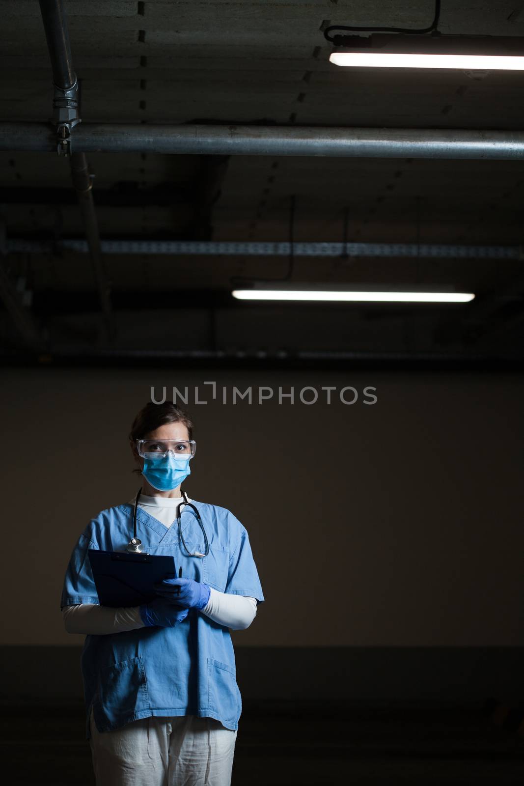 Portrait of frontline EMS key worker in dark hallway,in PPE blue uniform,protective face mask,goggles & gloves,holding clipboard patient form,worried stressed & overworked UK NHS ICU medical staff 