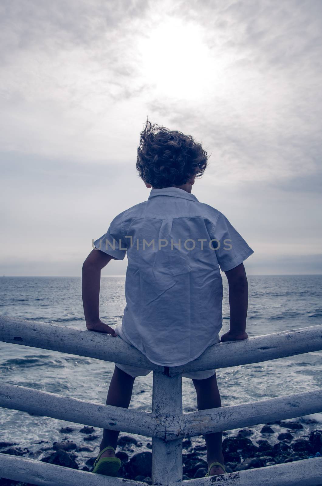 Boy with white clothes sitting on a fence facing the sea