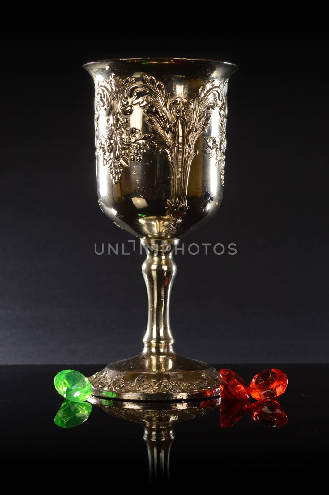 A silver cup with a few gemstones over a black reflective background.
