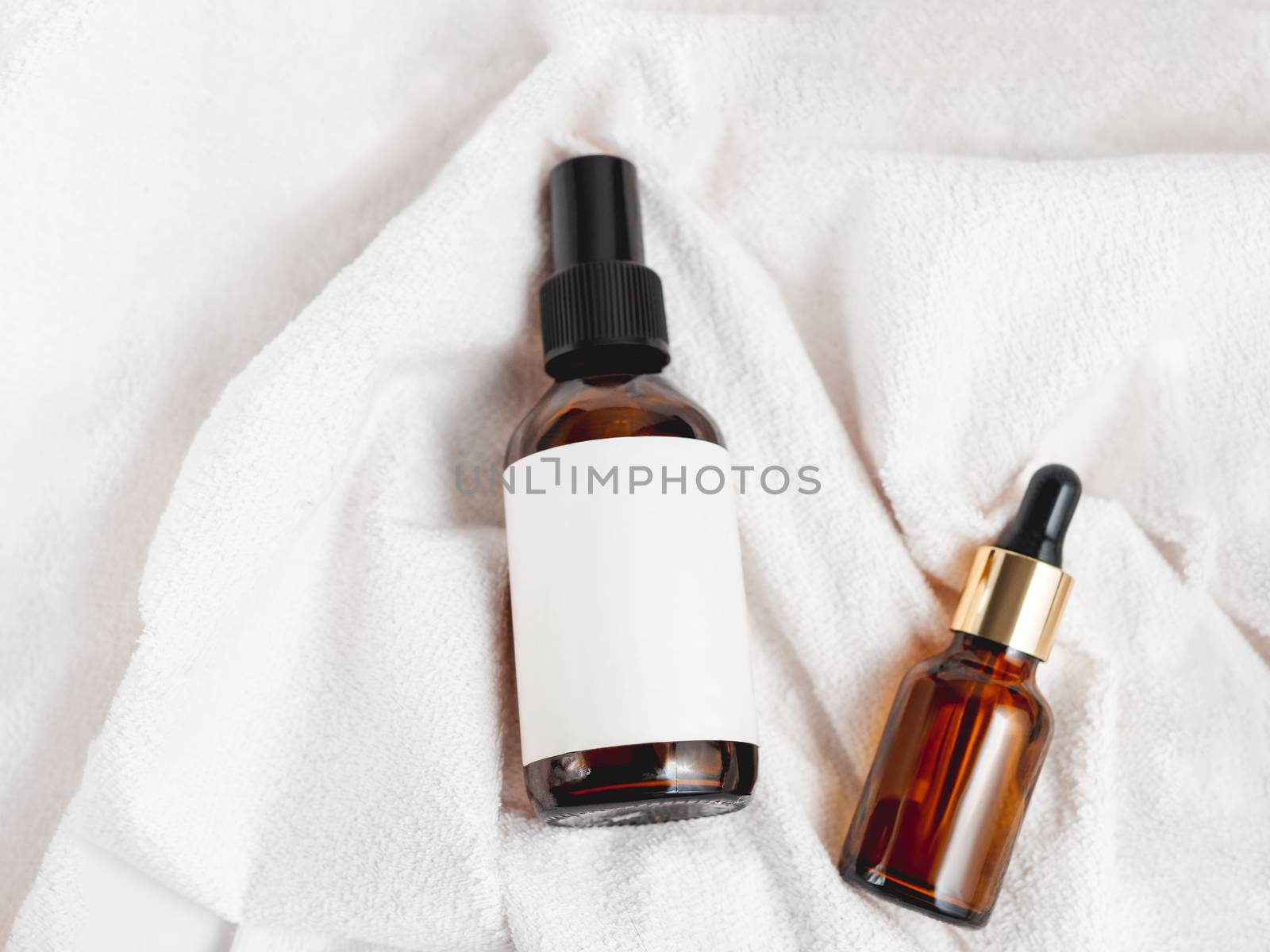 Top view on two glass bottles on white fluffy towel. Essential oil and cosmetic serum with clear labels on crumpled bathrobe